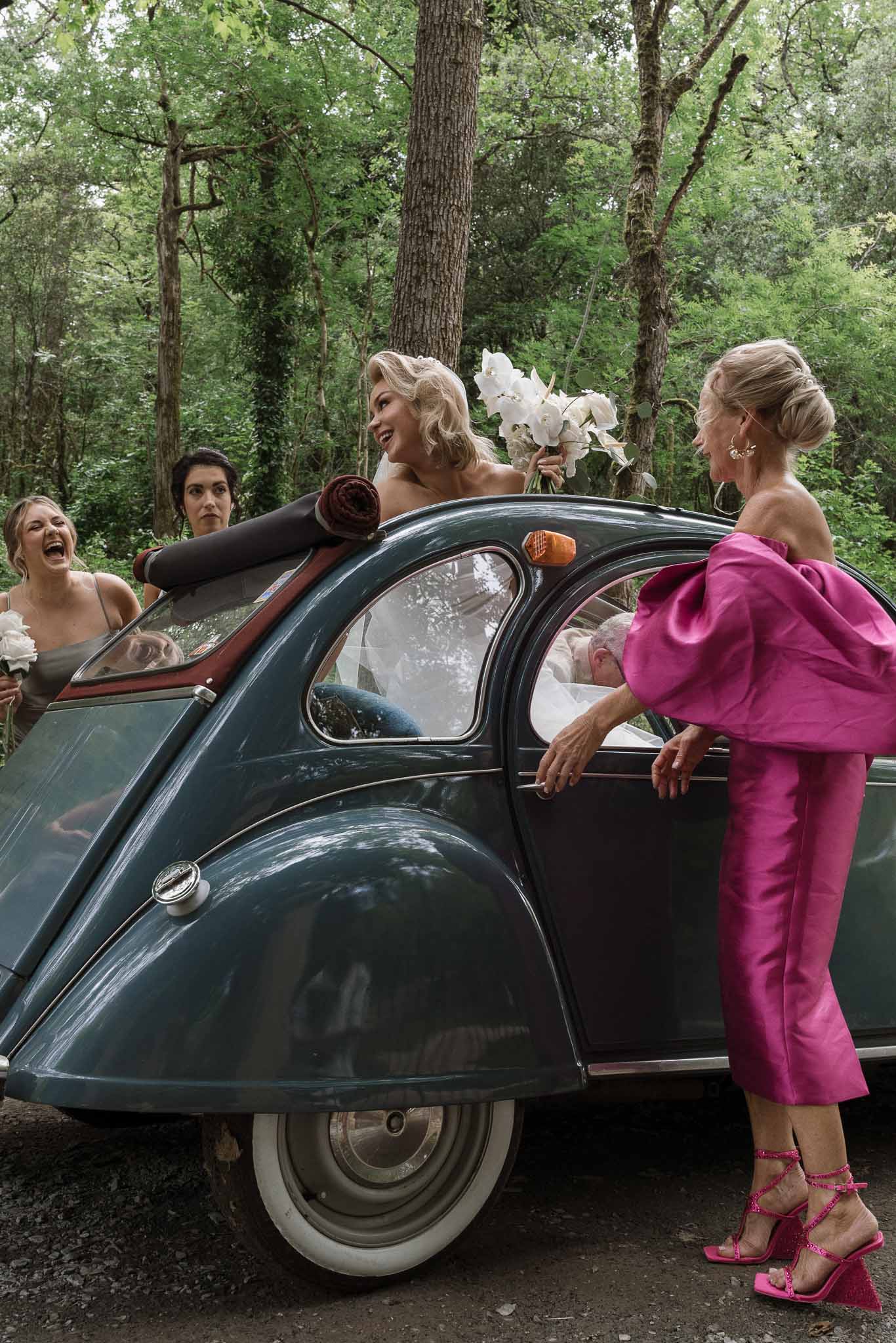 Bride and wedding party celebrating around vintage Citroën 2CV during wedding exit in wooded outdoor setting