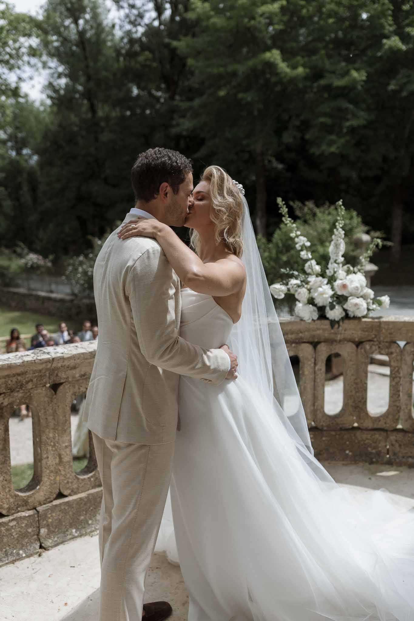 Bride and groom sharing first kiss during outdoor ceremony on stone terrace with garden backdrop