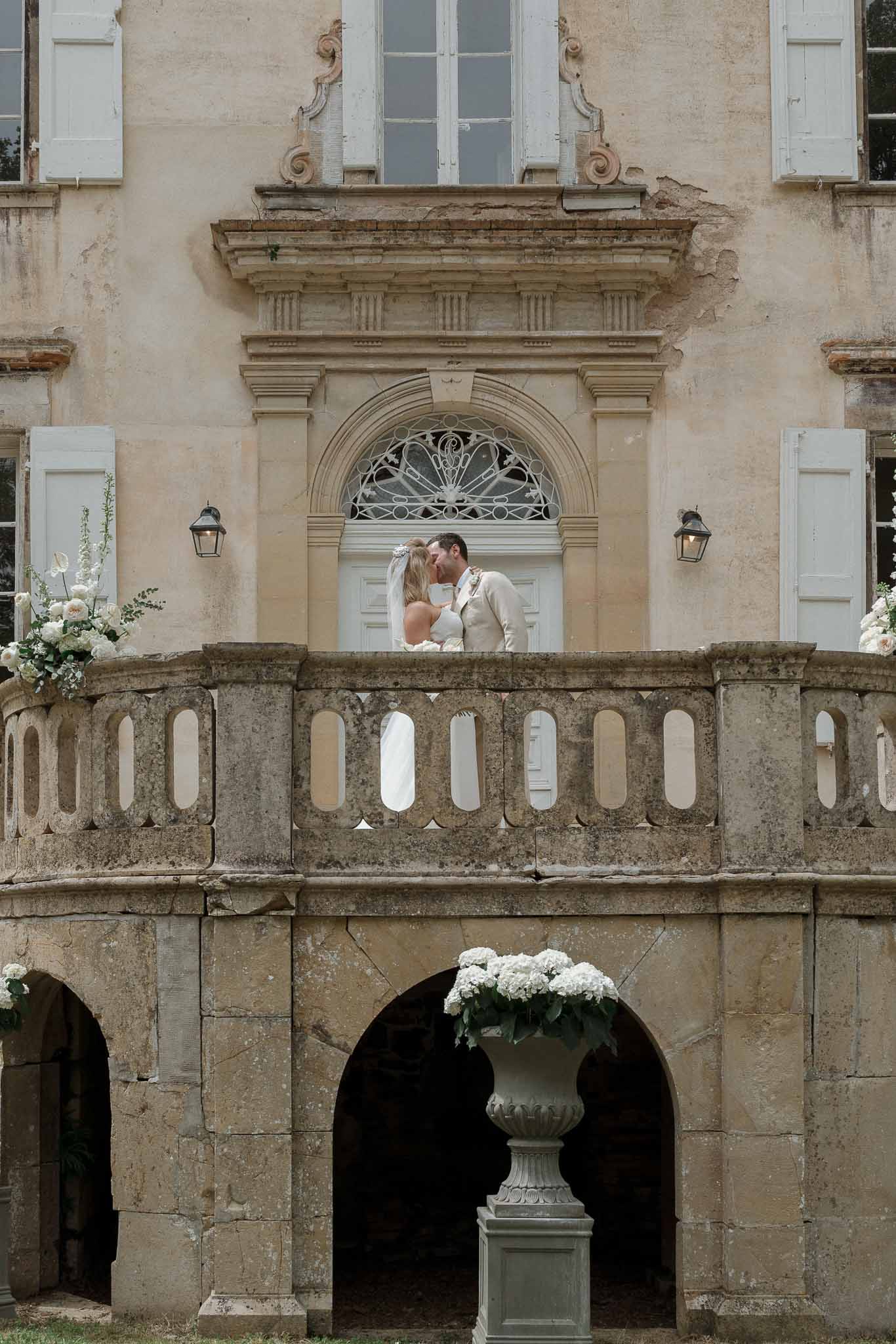 Bride and groom kissing on stone balcony of neoclassical mansion with white hydrangeas