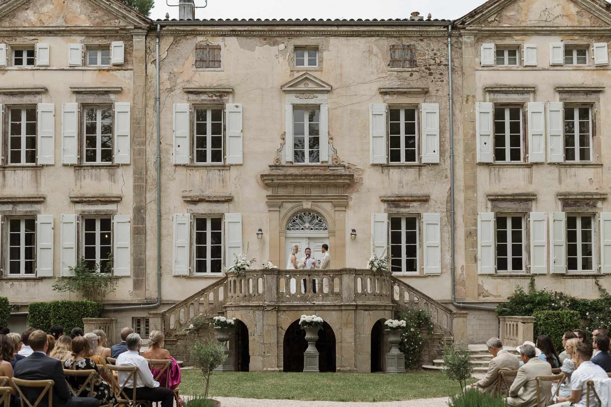Wedding ceremony on stone balcony of historic French château with guests seated on manicured lawn