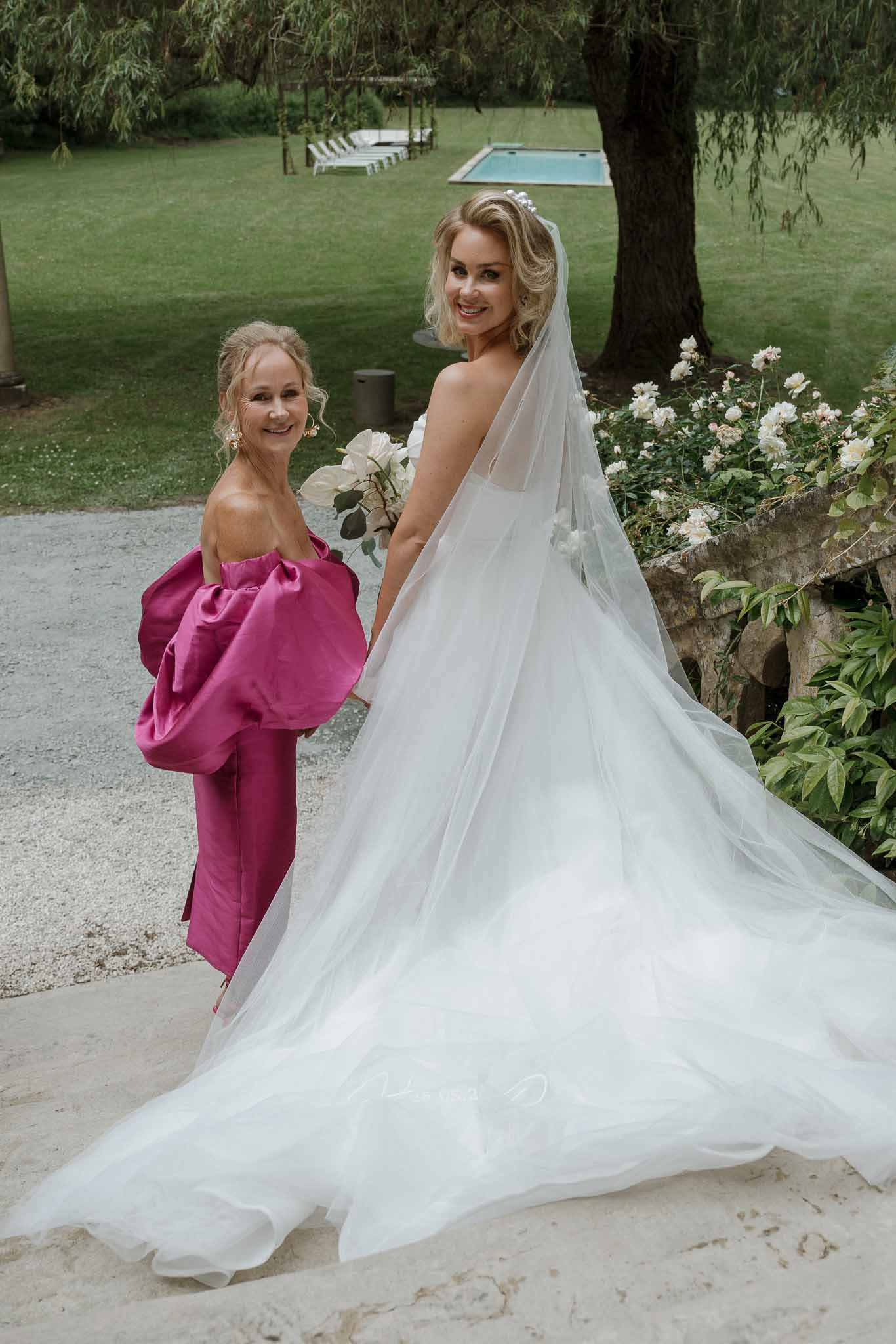 Bride and flower girl posing together in manicured garden estate with swimming pool in background