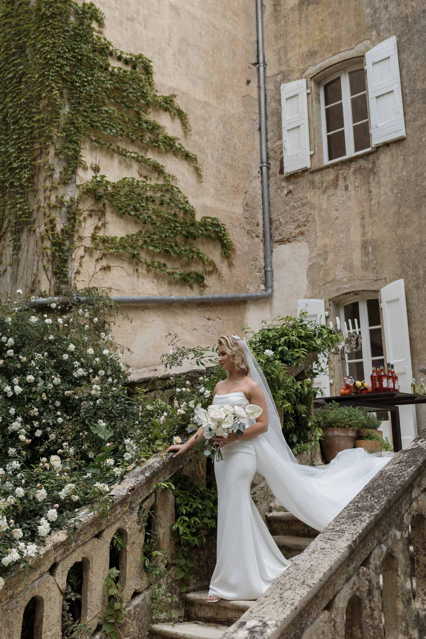 Bride in white gown with veil and bouquet posing in historic stone courtyard with ivy-covered walls