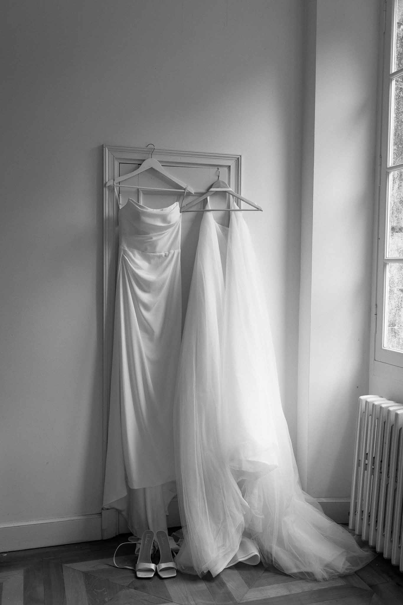 Two wedding dresses hanging on mirror with bridal shoes in bright indoor room