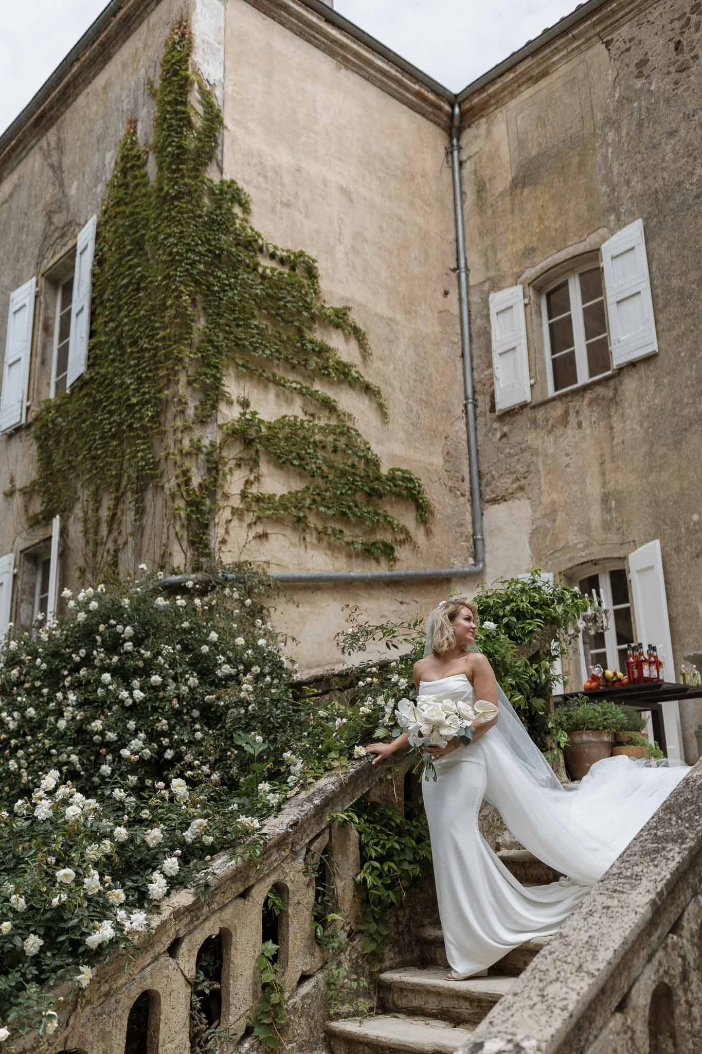 Bride in ivory gown with white bouquet standing on stone steps at historic courtyard venue