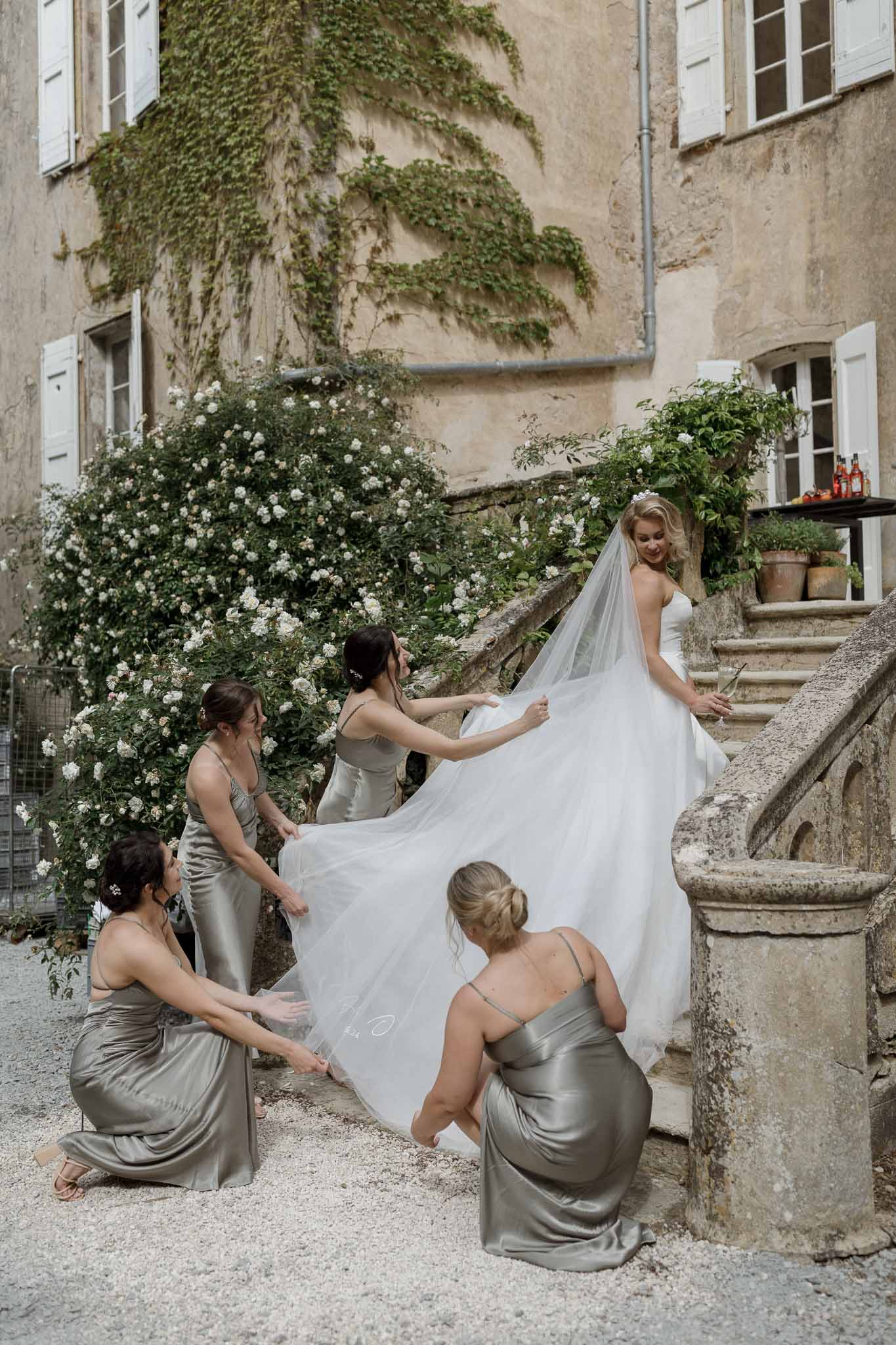 Bride with four bridesmaids arranging dress and train in stone courtyard with ivy-covered European building