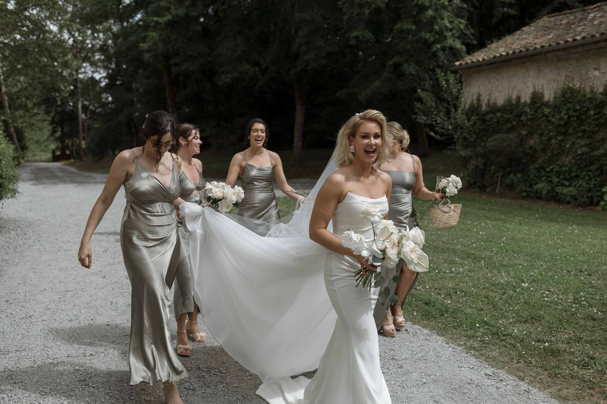 Bride and bridesmaids walking along tree-lined gravel driveway at outdoor estate wedding venue