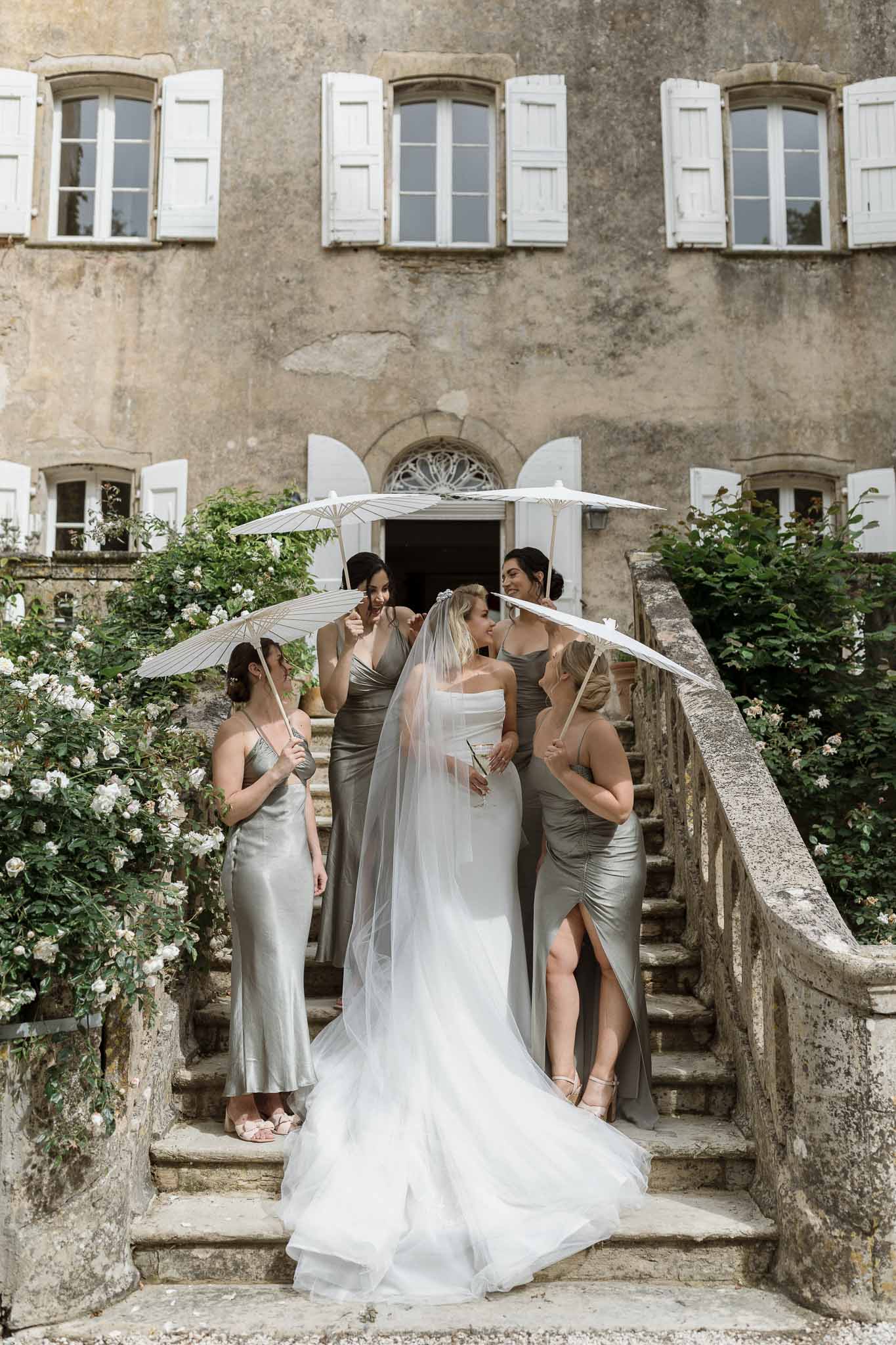Bride and bridesmaids with cream umbrellas on stone stairs at European villa during getting ready