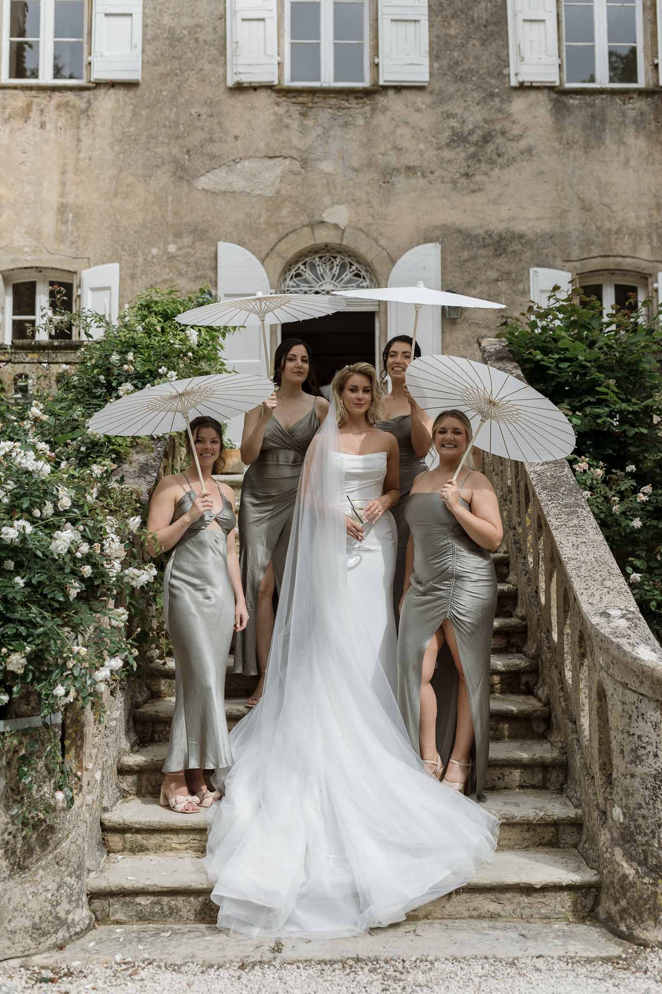 Bride and bridesmaids with parasols on stone staircase at historic estate