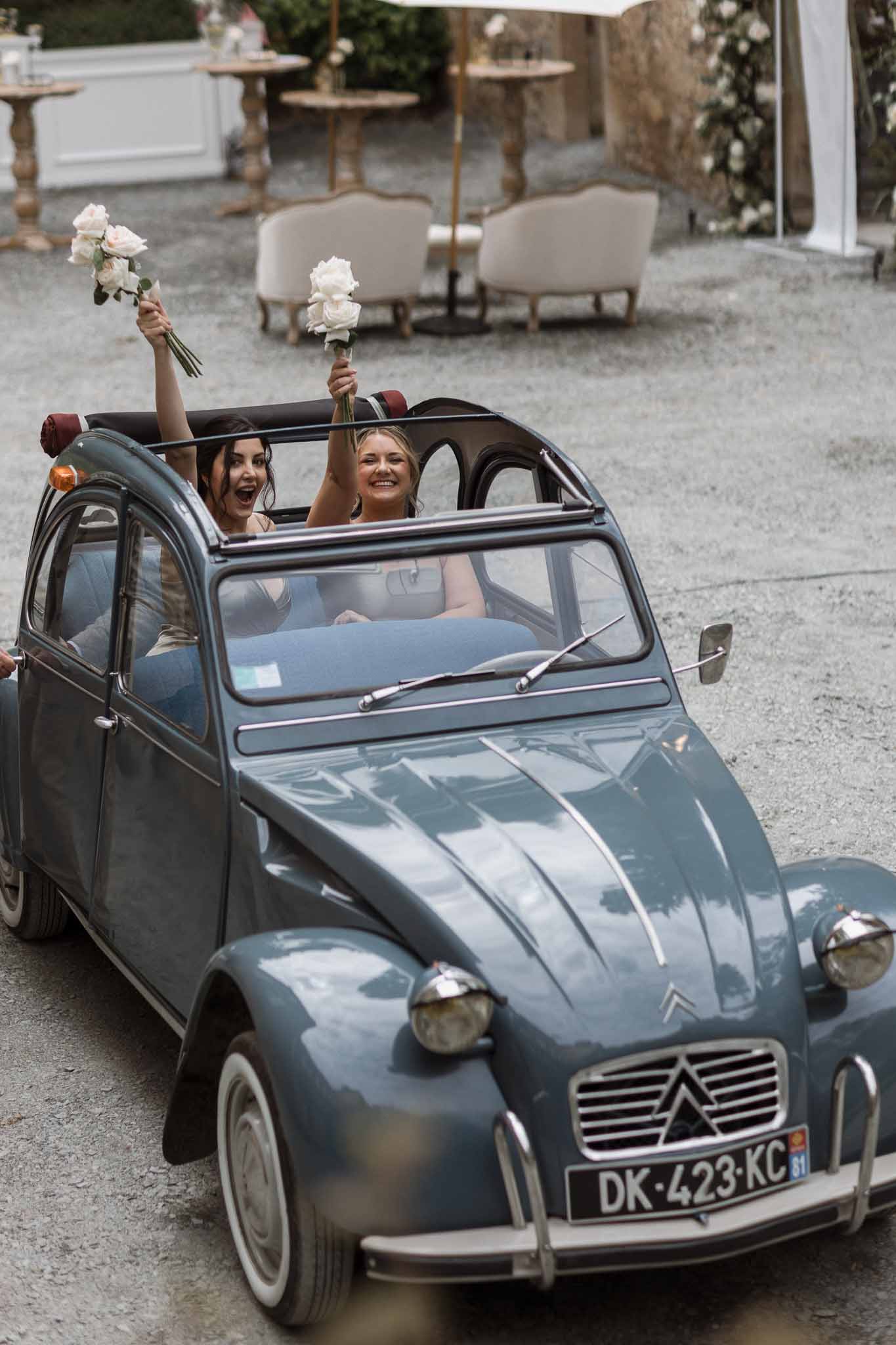 Bride and bridesmaid in vintage Citroën 2CV with bouquets at outdoor courtyard wedding reception