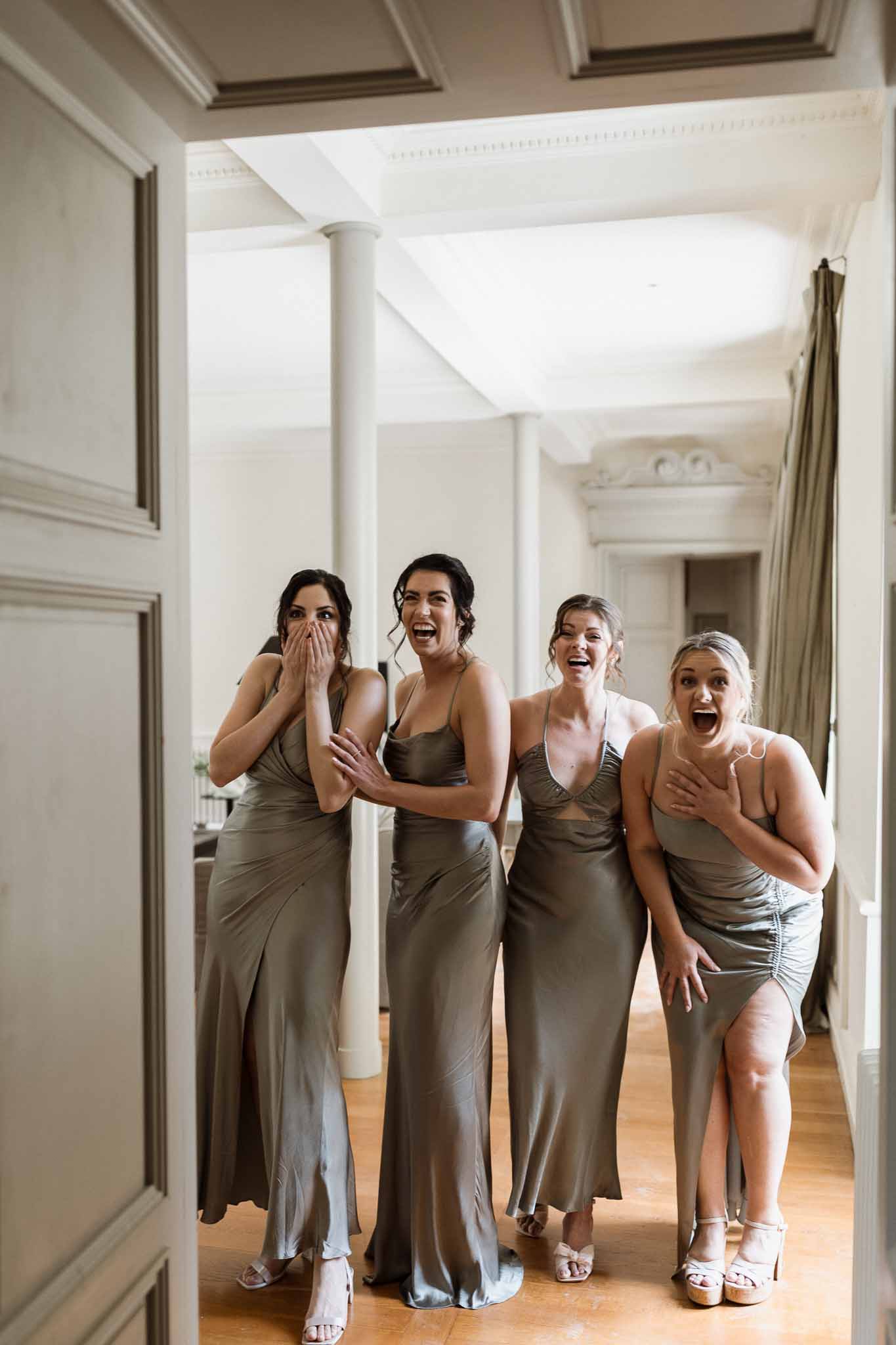 Four bridesmaids in sage green dresses laughing together in classical hallway with white pillars