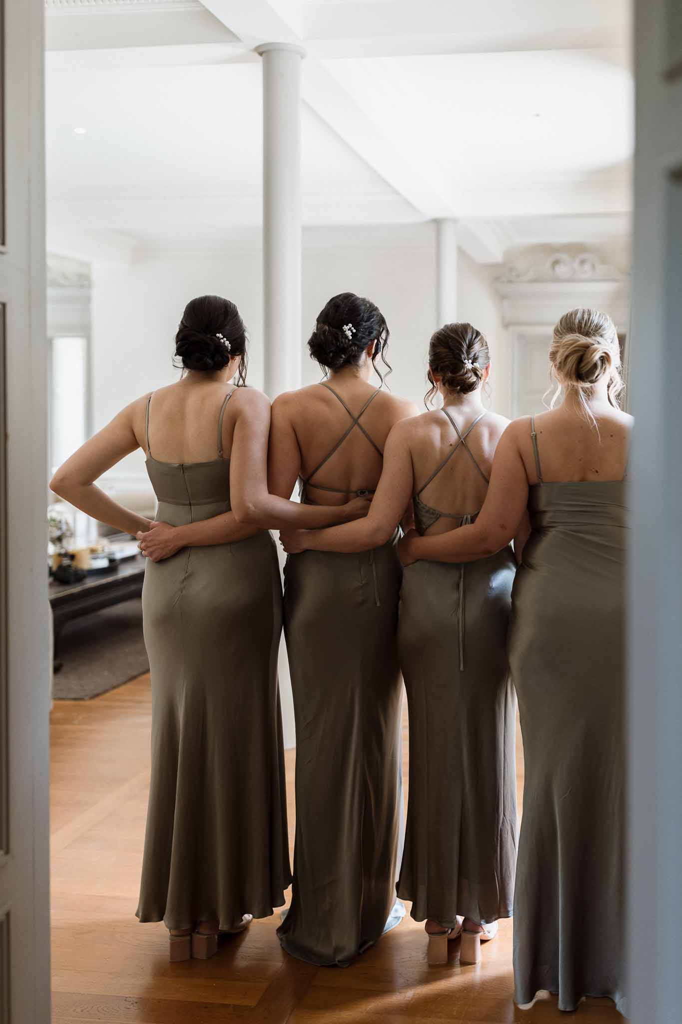 Four bridesmaids in sage green dresses standing together in modern interior space with white columns