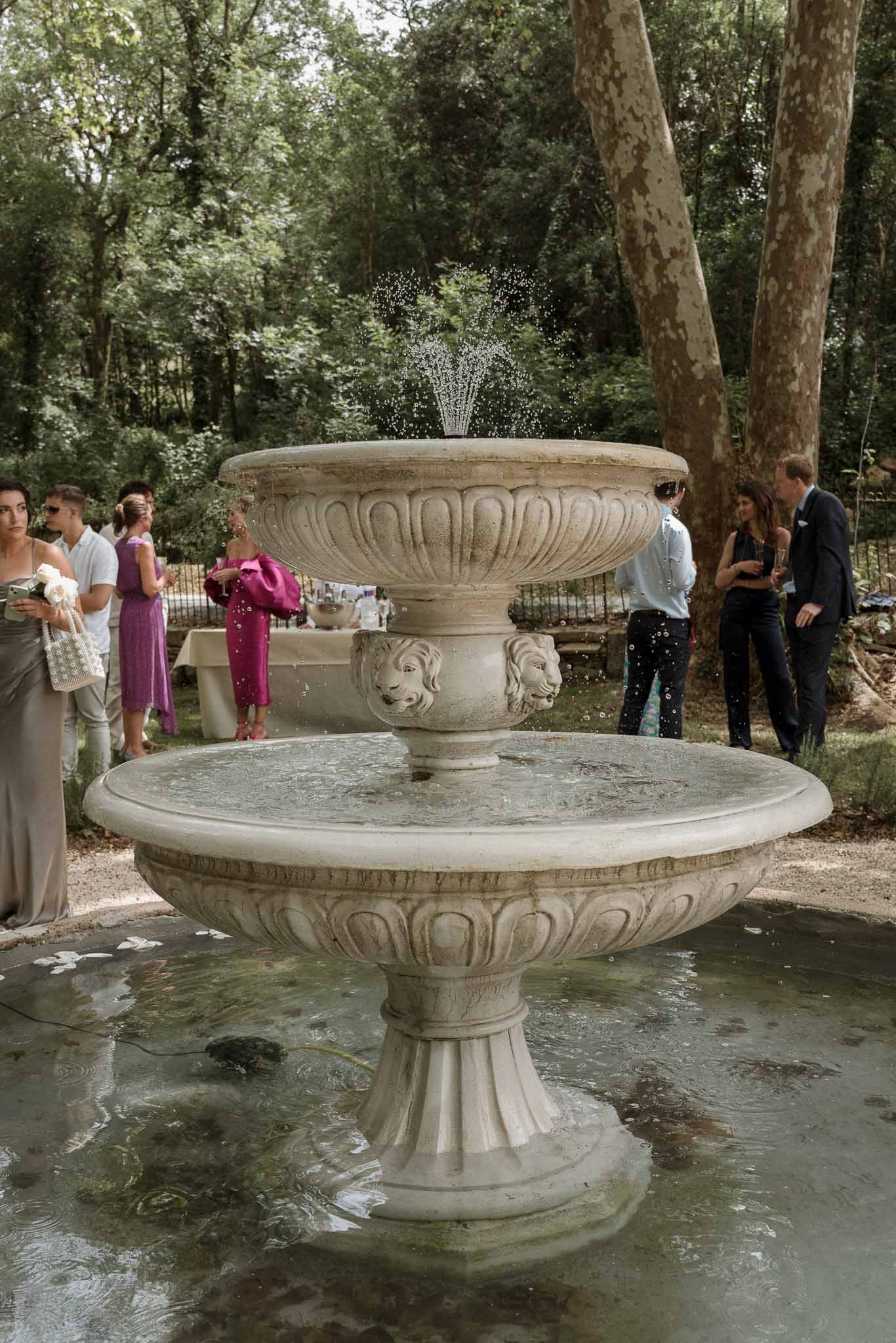 Wedding guests gathering around ornate stone fountain during cocktail hour in woodland garden setting