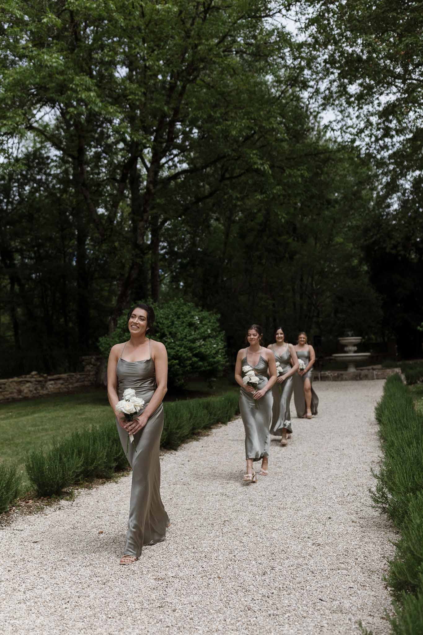Bridesmaids in sage green dresses walking processional through formal garden with lavender hedging