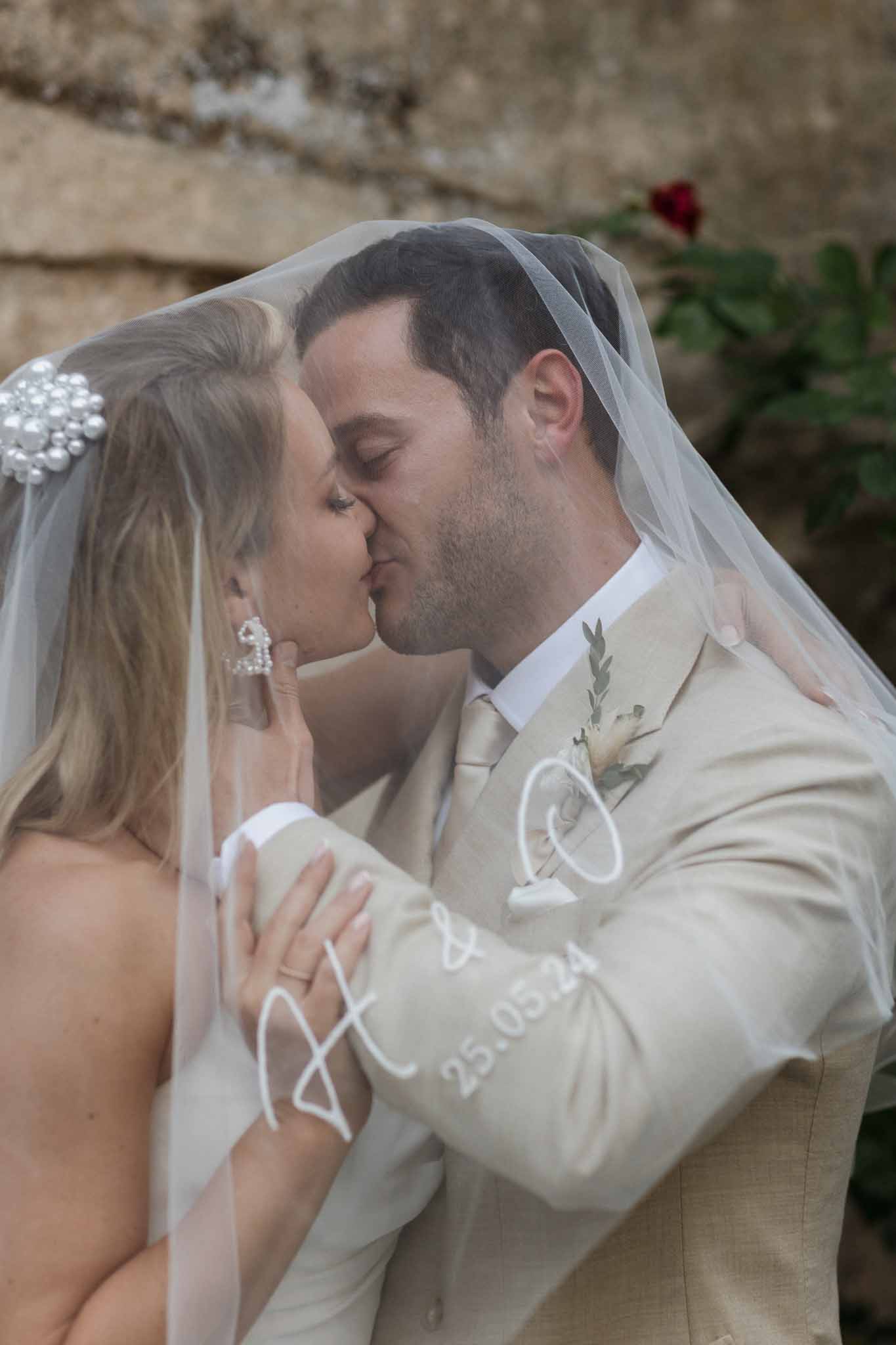 Bride and groom first kiss during wedding ceremony with tulle veil and stone wall backdrop
