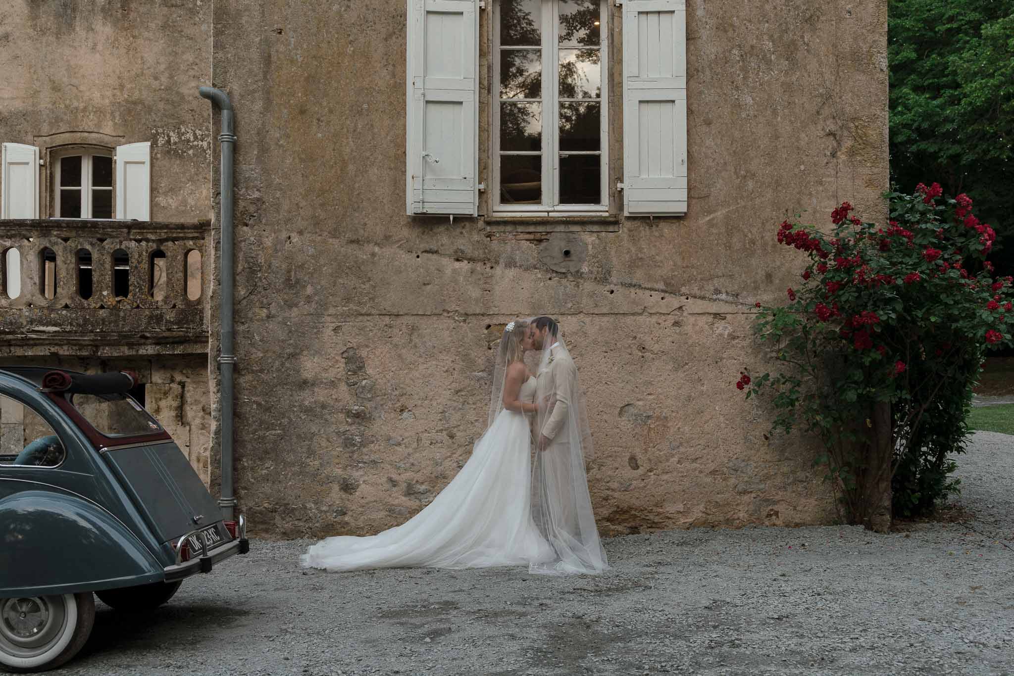 Bride and groom kissing in stone courtyard with vintage car and climbing roses