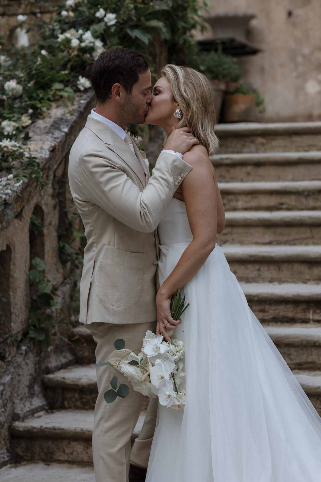 Bride and groom kissing on stone staircase in outdoor courtyard with ivy-covered walls