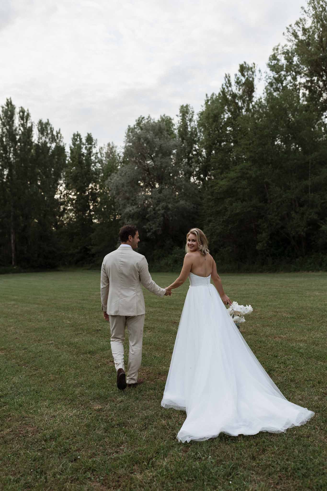 Newlywed couple walking hand-in-hand across grassy field with poplar trees