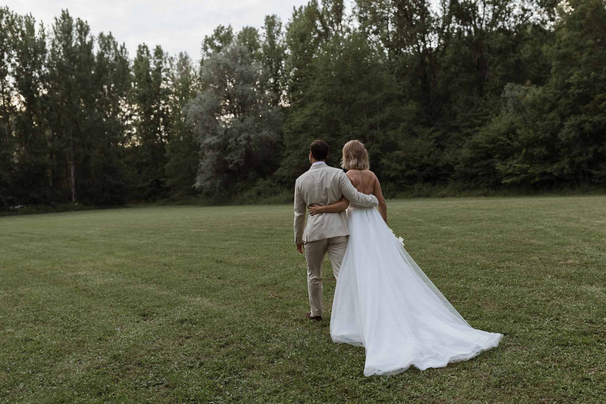 Bride and groom walking together across meadow with trees in background