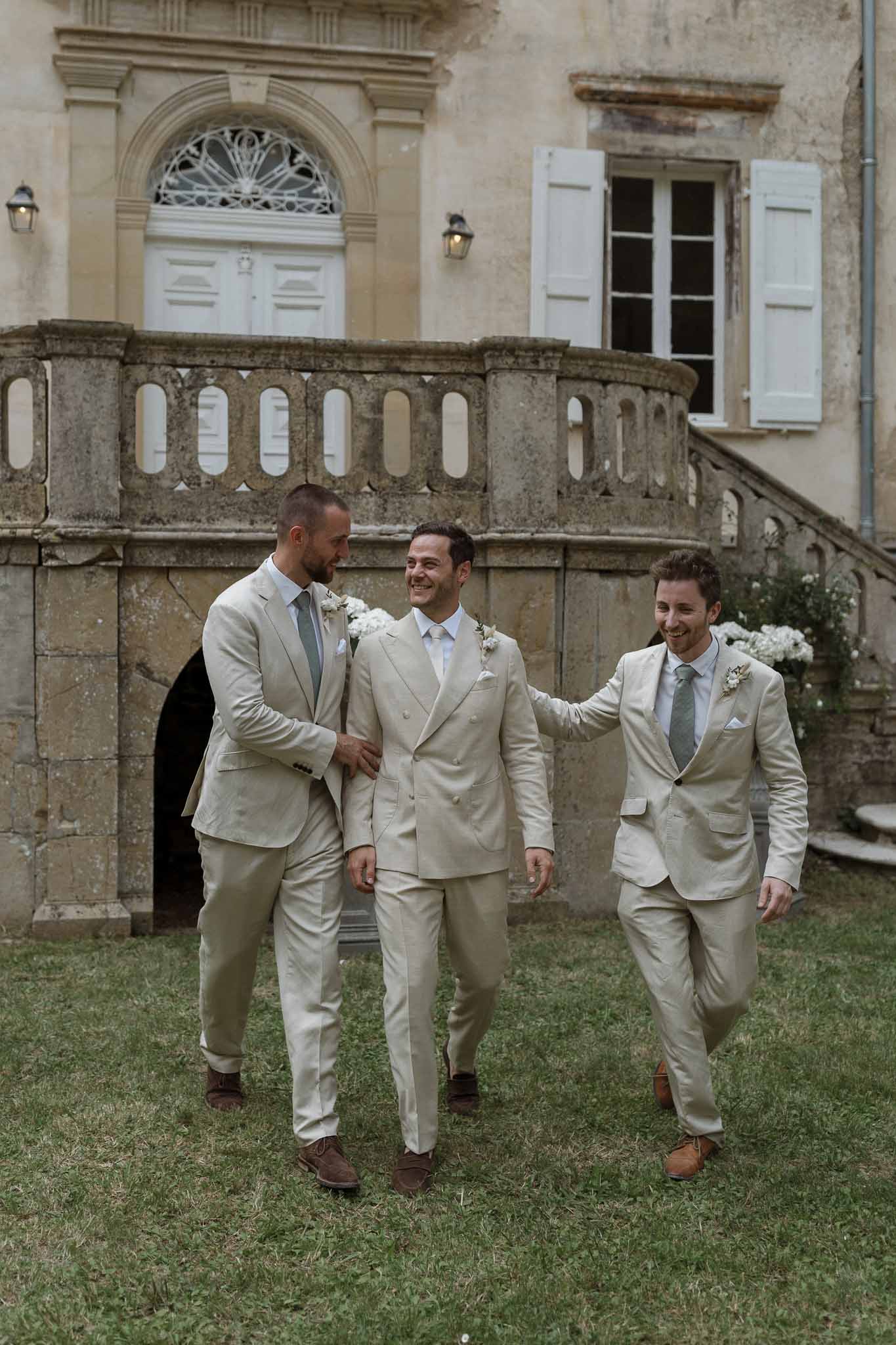 Three groomsmen in cream linen suits walking across lawn at historic French château