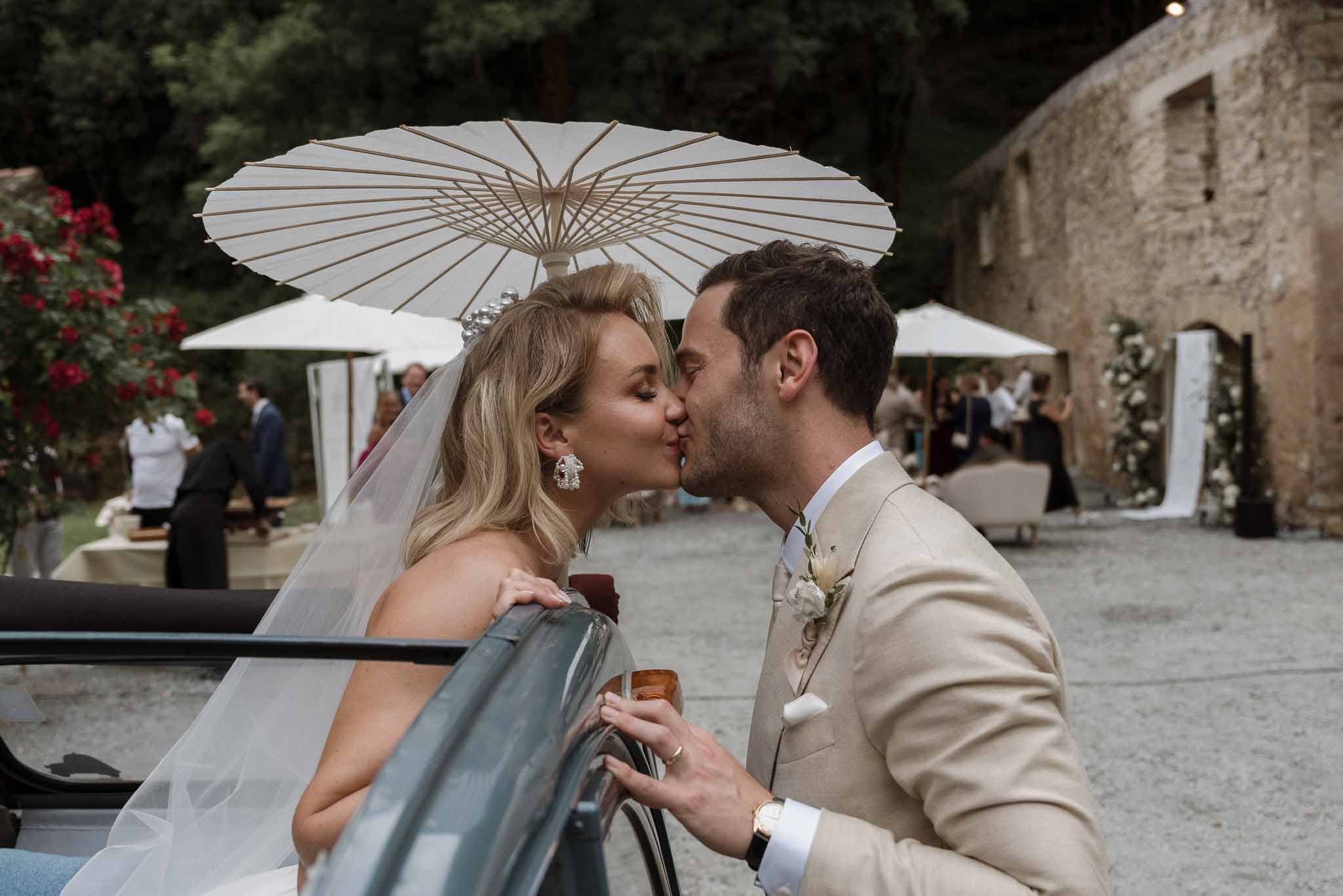 Bride and groom kissing under parasol in historic stone courtyard during wedding reception