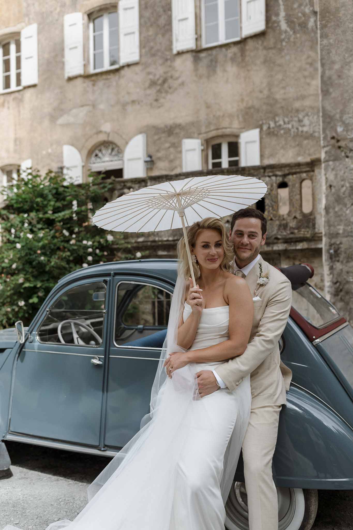 Bride and groom posing with vintage car in stone courtyard with parasol
