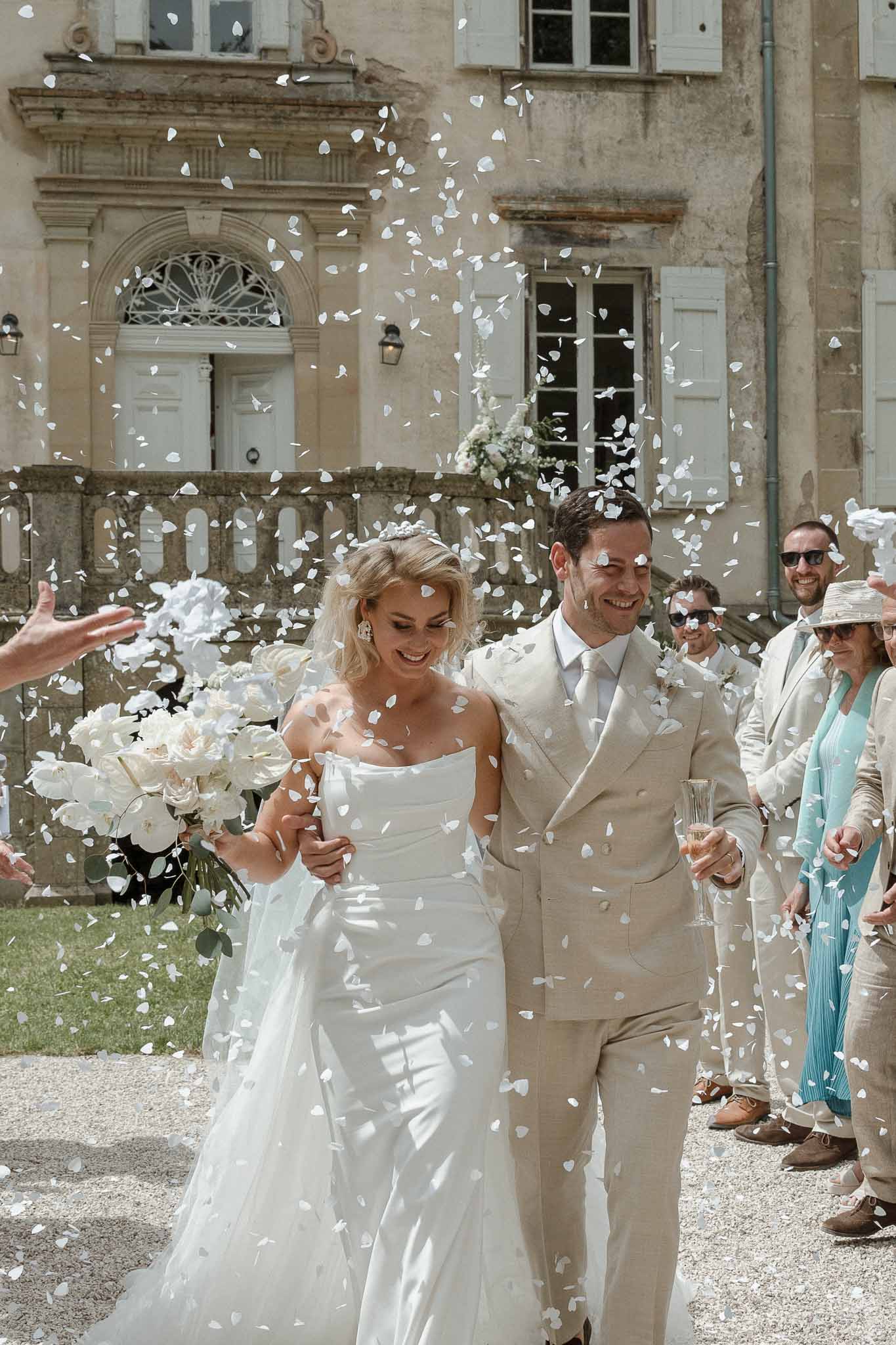 Bride and groom walking through petal toss in European stone courtyard with guests celebrating