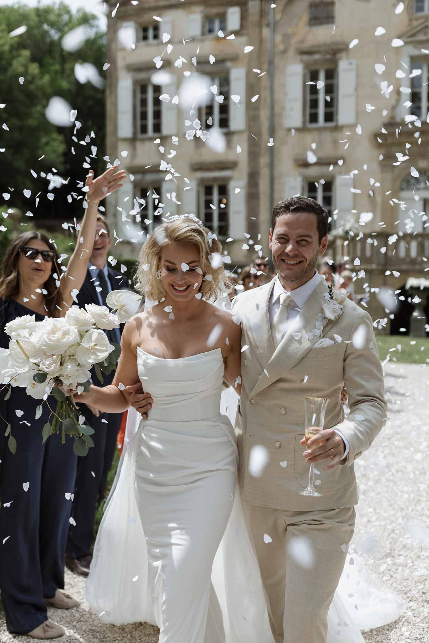 Newlyweds walking through petal toss in château courtyard with wedding guests
