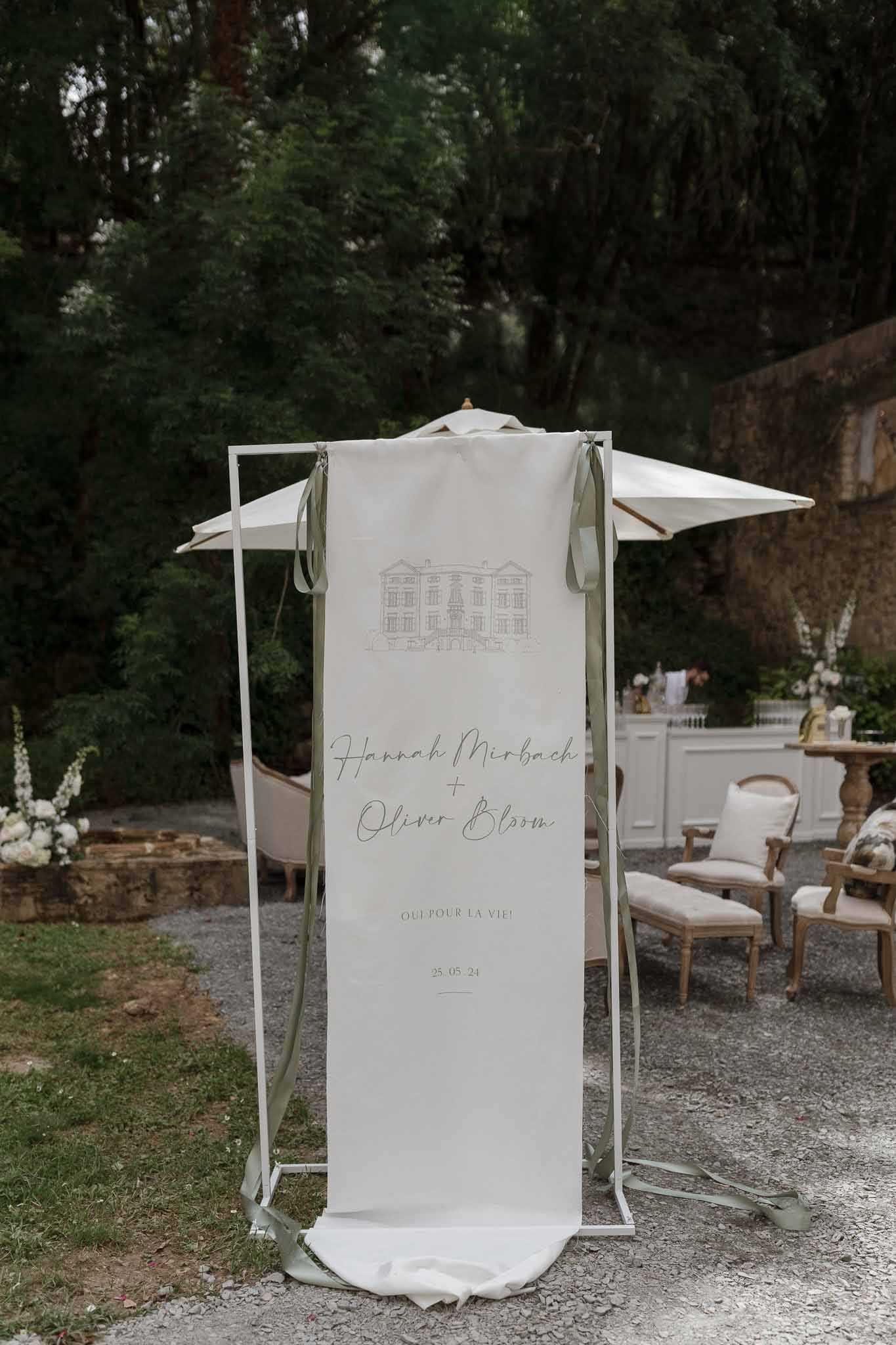 Wedding welcome banner with couple names and date hanging from white pergola in garden courtyard