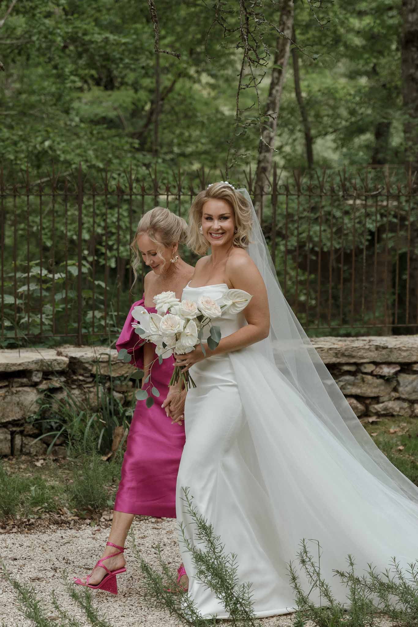 Bride with companion in fuchsia dress standing together in garden setting with wrought-iron fence
