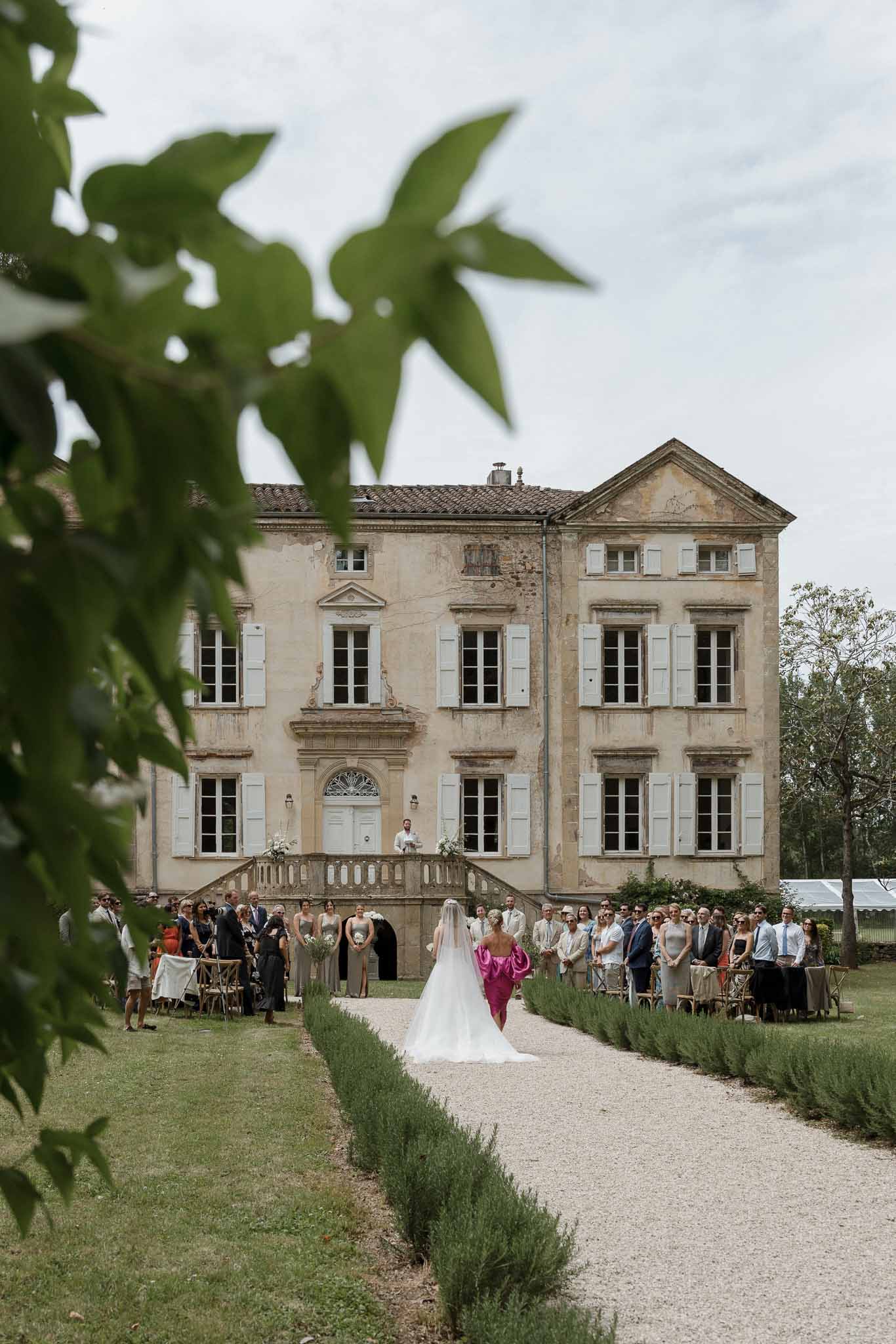 Bride walking down aisle during outdoor ceremony at historic stone château with guests seated in garden