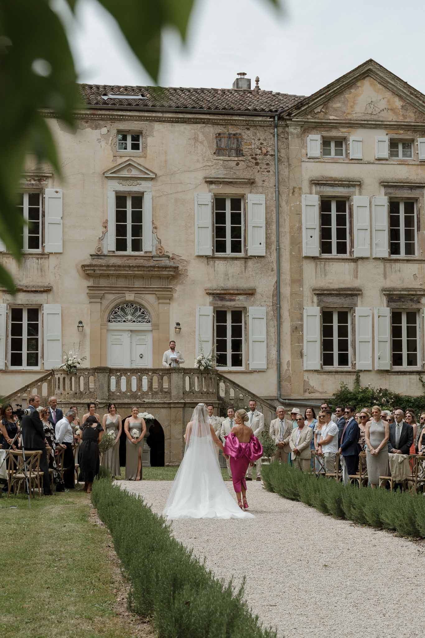 Bride walking down aisle during outdoor ceremony at French château with guests in wooden chairs