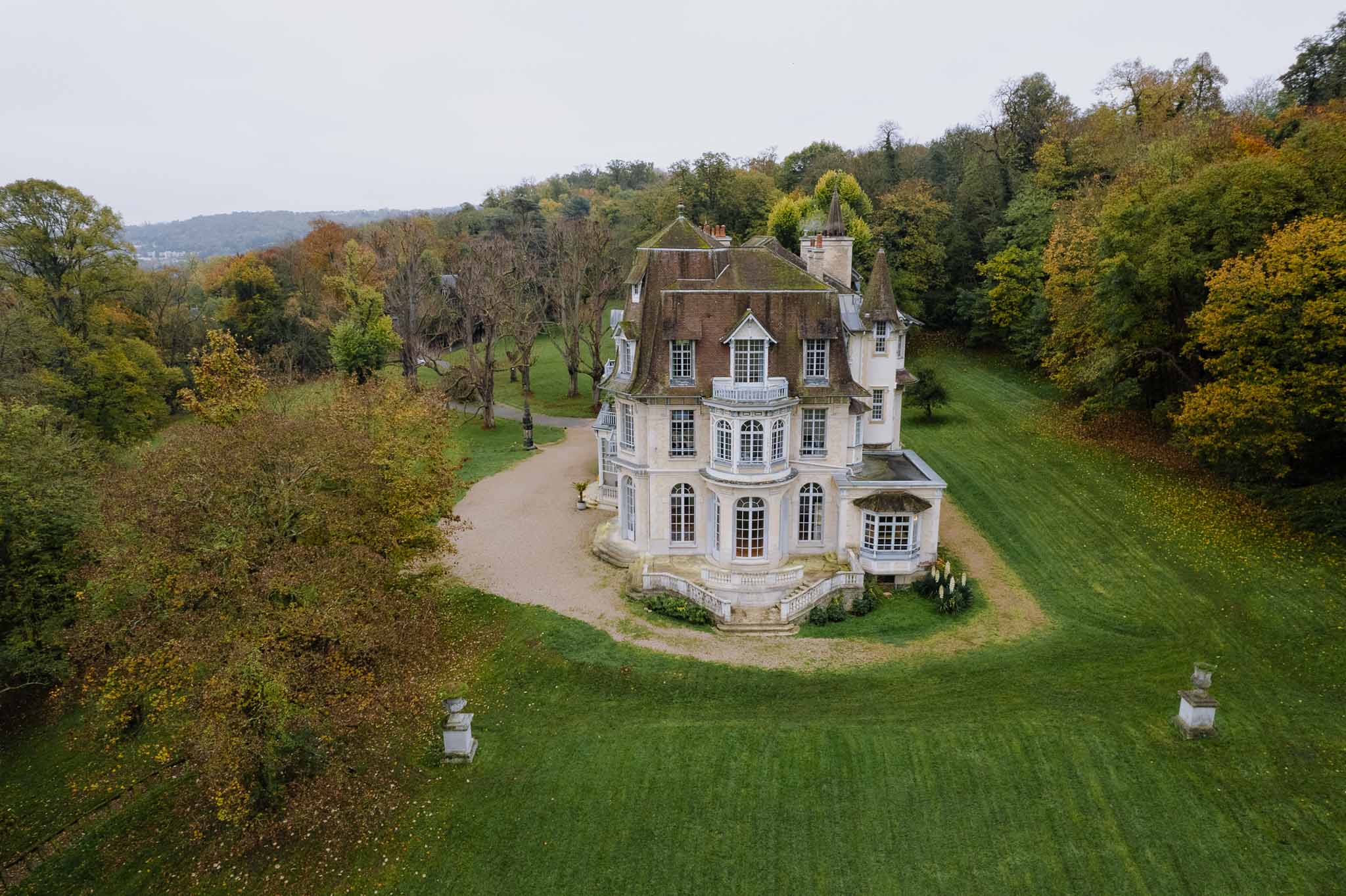 Aerial view of historic château with turrets and formal grounds surrounded by autumn woodland