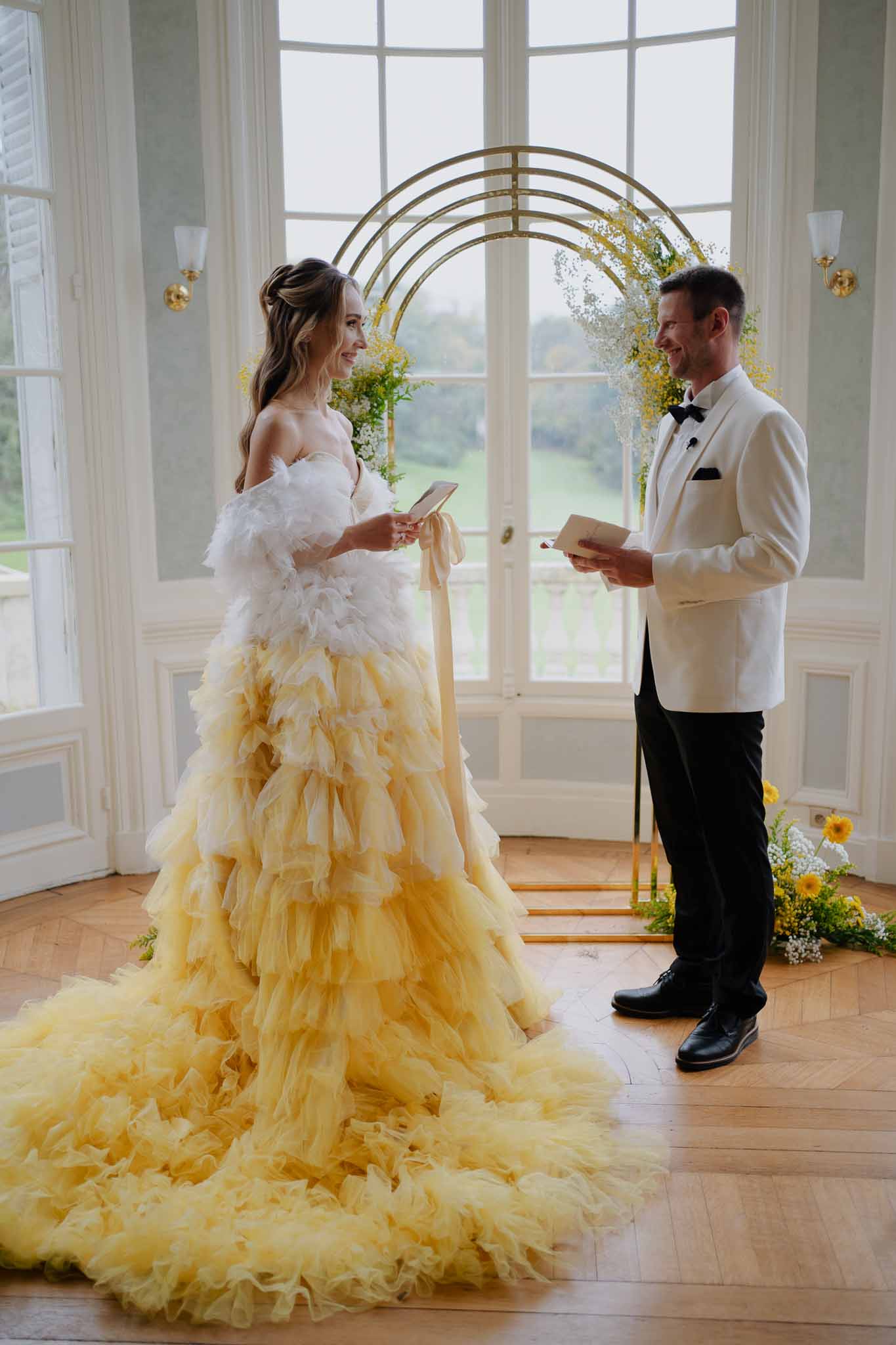 Bride and groom exchanging vows under floral arch during indoor wedding ceremony with floor-to-ceiling windows