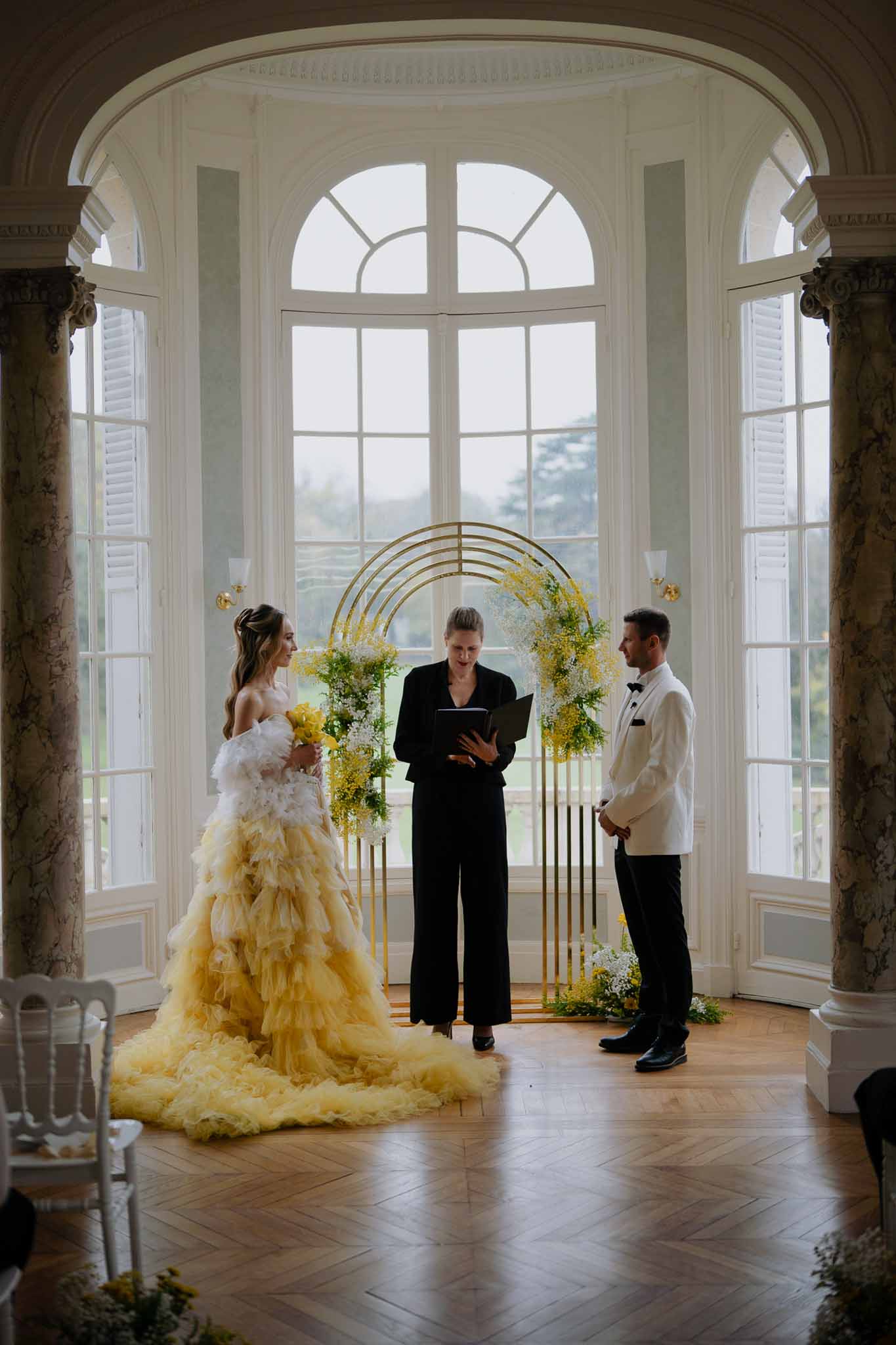 Wedding ceremony in neoclassical ballroom with bride, groom, and officiant under gold floral arch