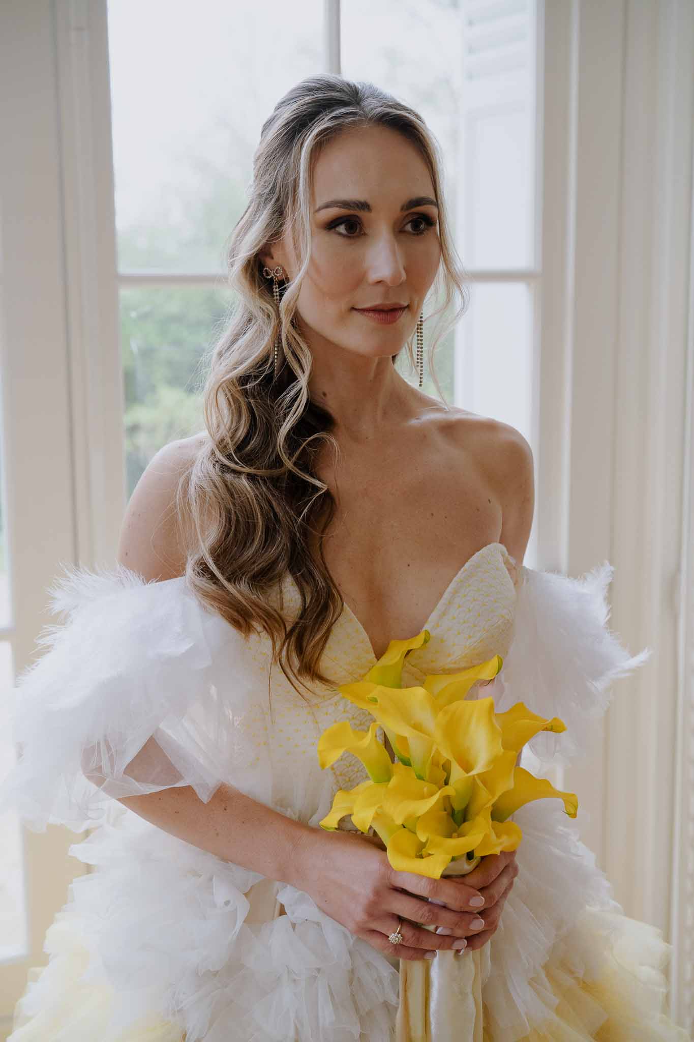Bride in off-shoulder gown holding yellow lily bouquet by window during indoor portrait session