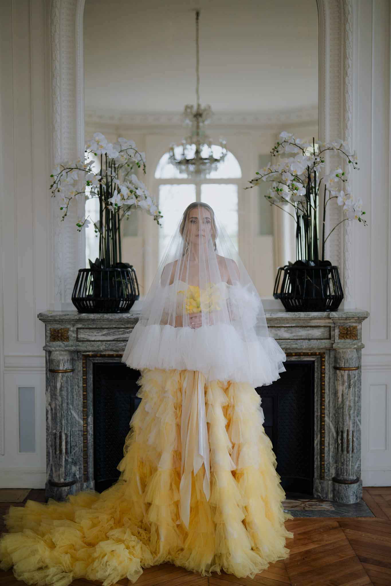 Bride in ivory and pale yellow gown standing by stone fireplace in elegant formal room