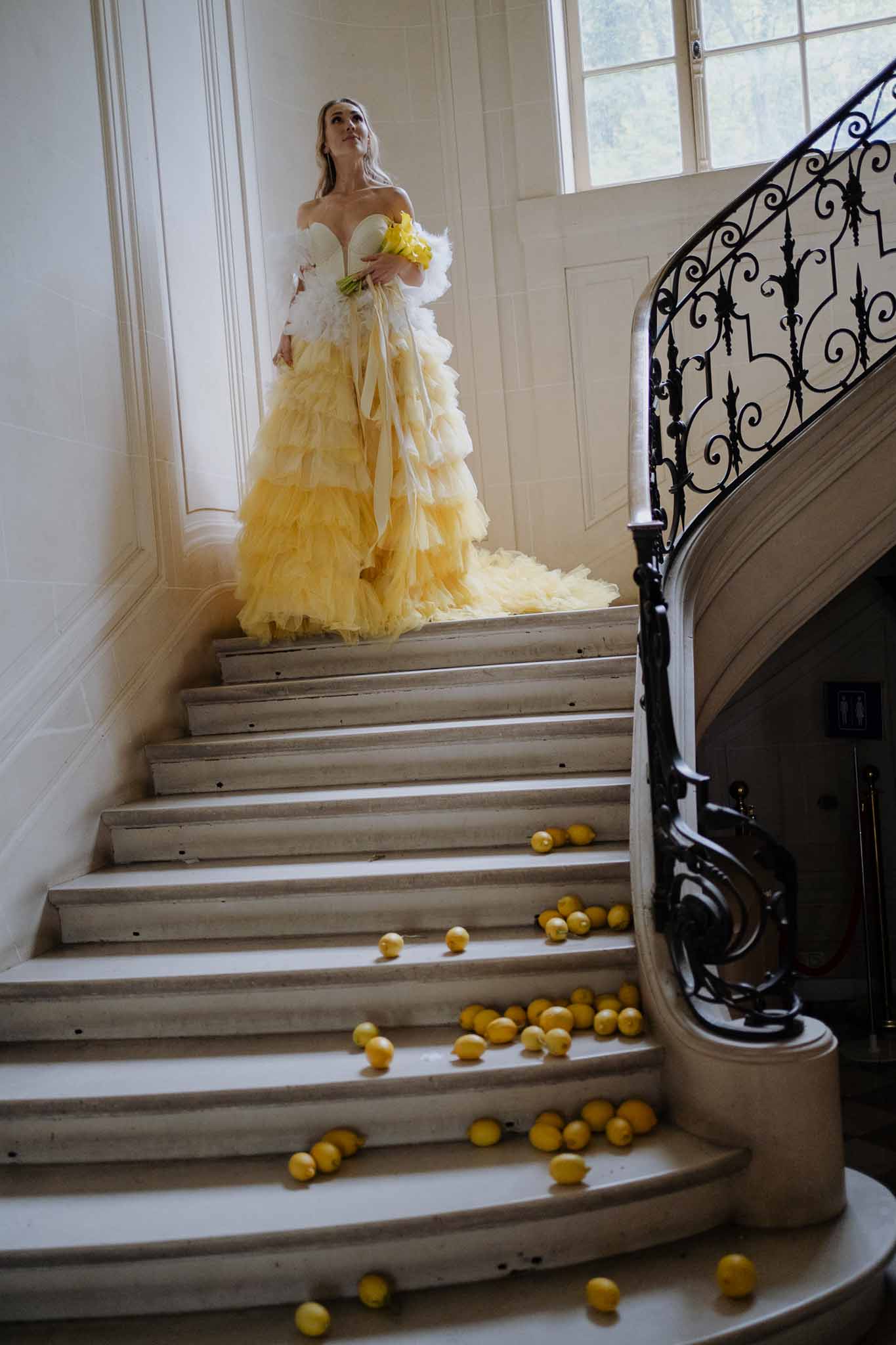 Bride in cream and yellow gown posing on grand staircase with lemon decorations in elegant interior setting