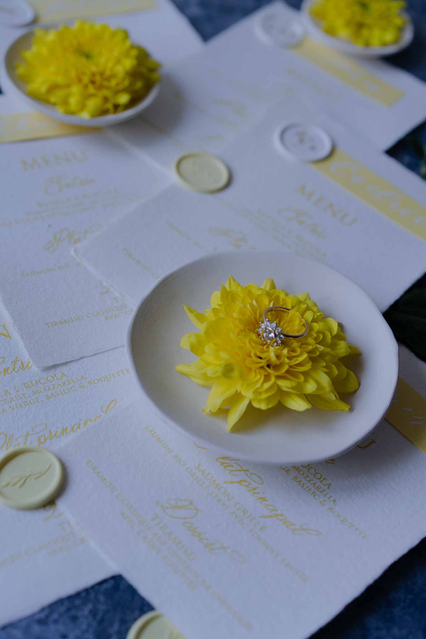 Yellow dahlia and engagement ring detail on white menu card with gold text at wedding reception table setting