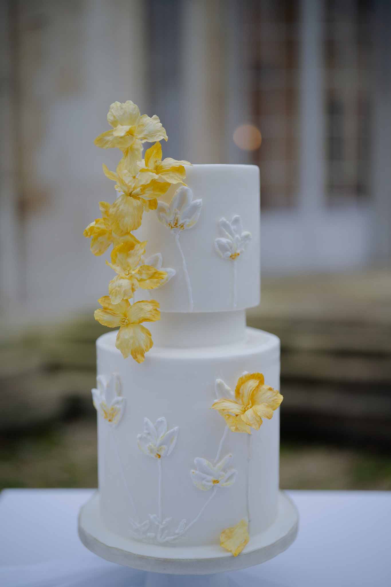 Two-tier white wedding cake with painted flowers and yellow iris blooms at outdoor venue