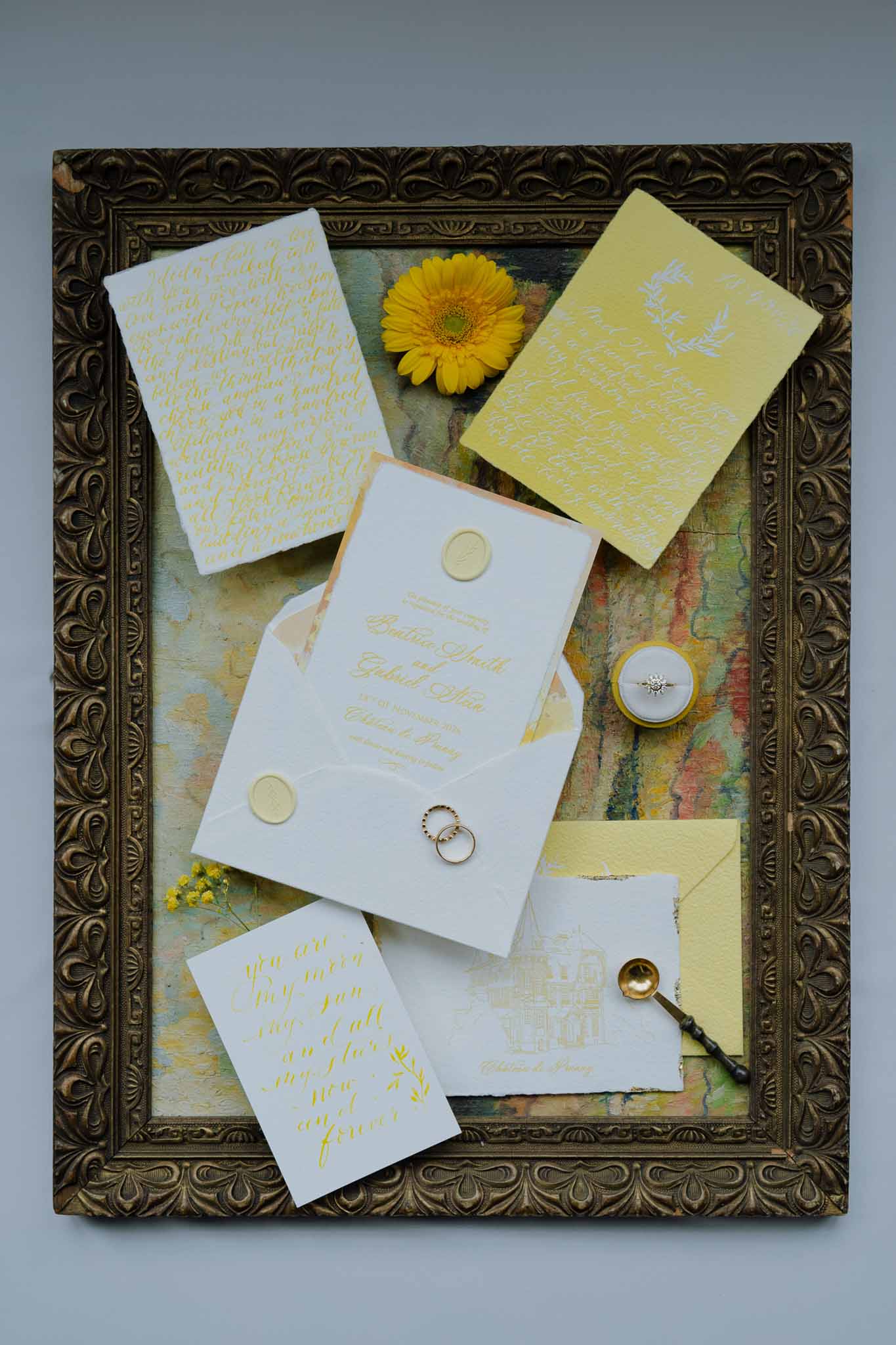 Wedding stationery flat lay with gold calligraphy invitations, rings, and yellow gerbera daisy in ornate frame