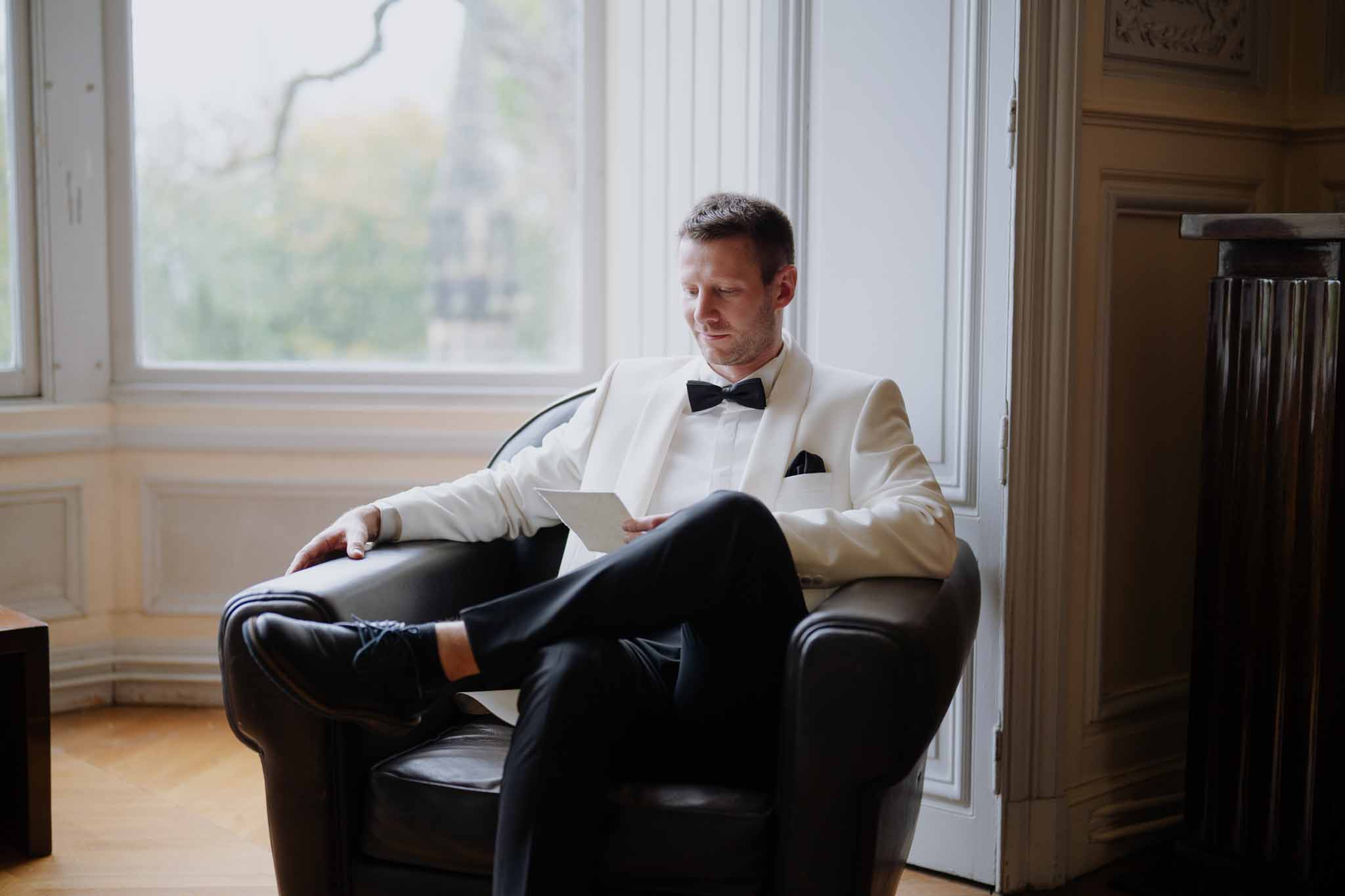 Groom in cream dinner jacket reviewing vows on tablet in elegant interior with tall windows and parquet flooring