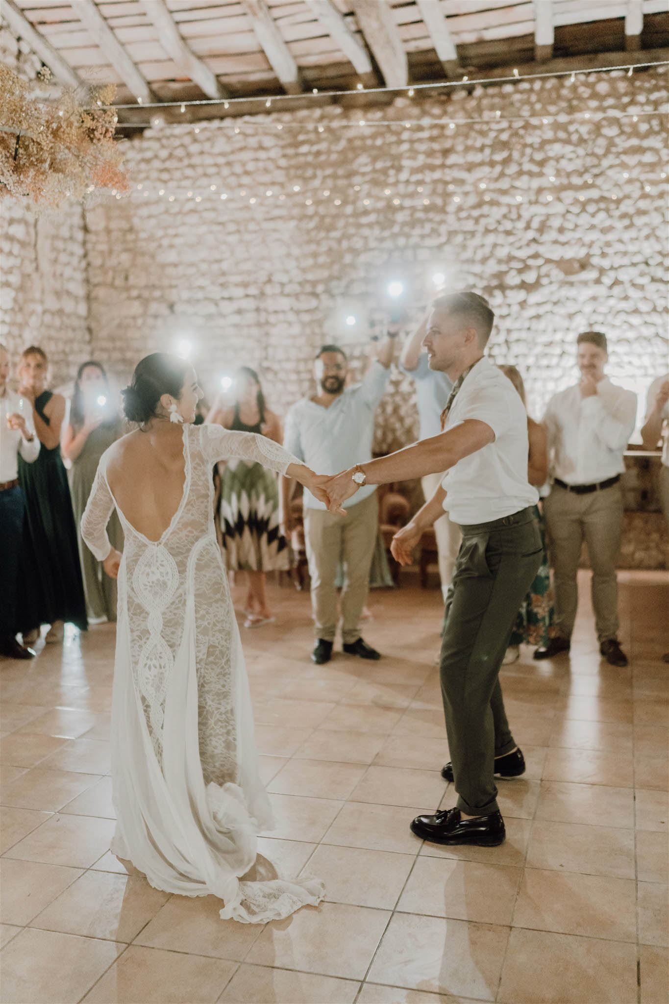 First dance in stone-walled venue with fairy lights guests holding phone flashlights bride in lace open-back gown