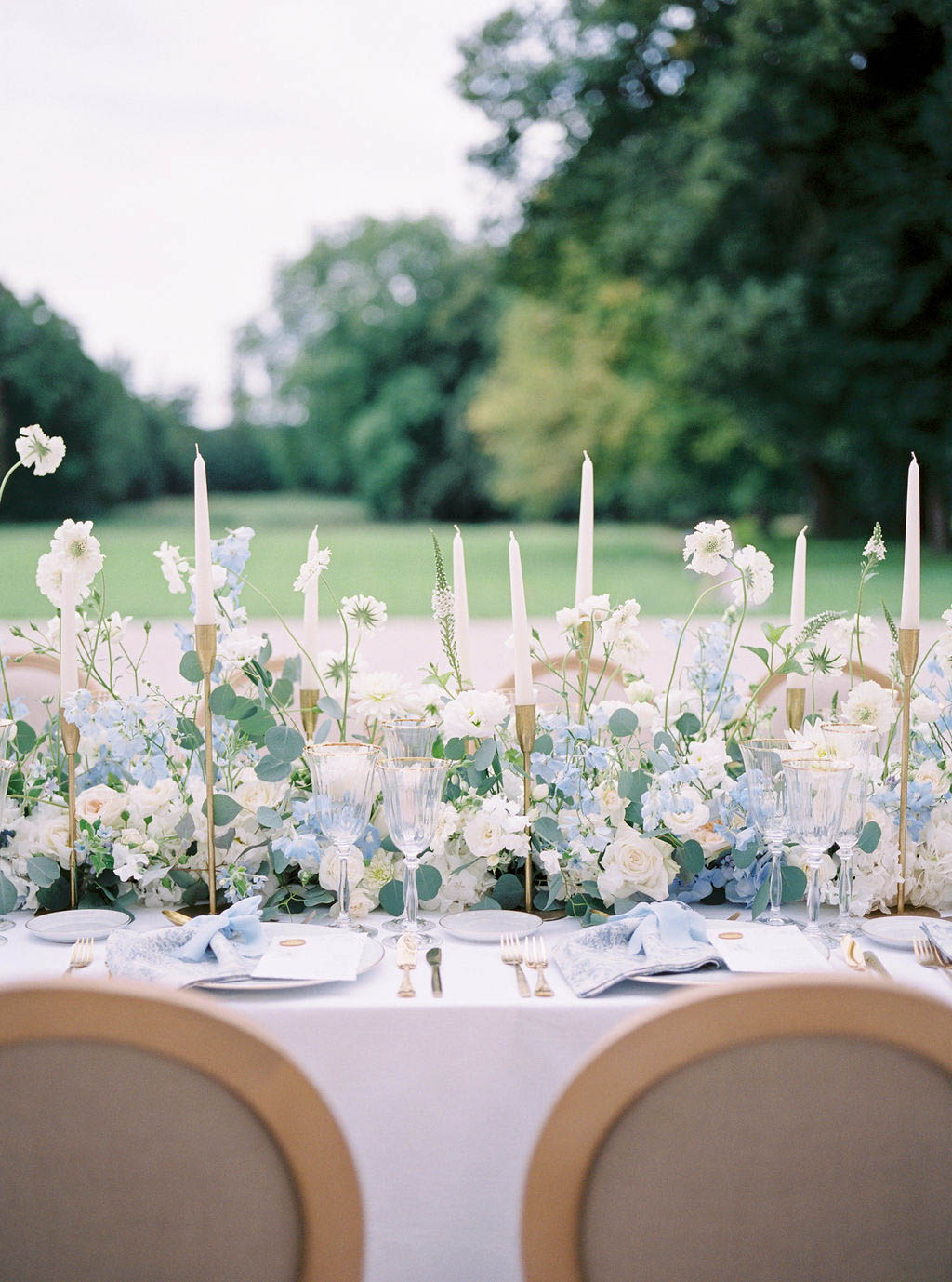 Reception tablescape with powder blue hydrangeas, white roses, brass candle holders, gold flatware, and crystal glasses