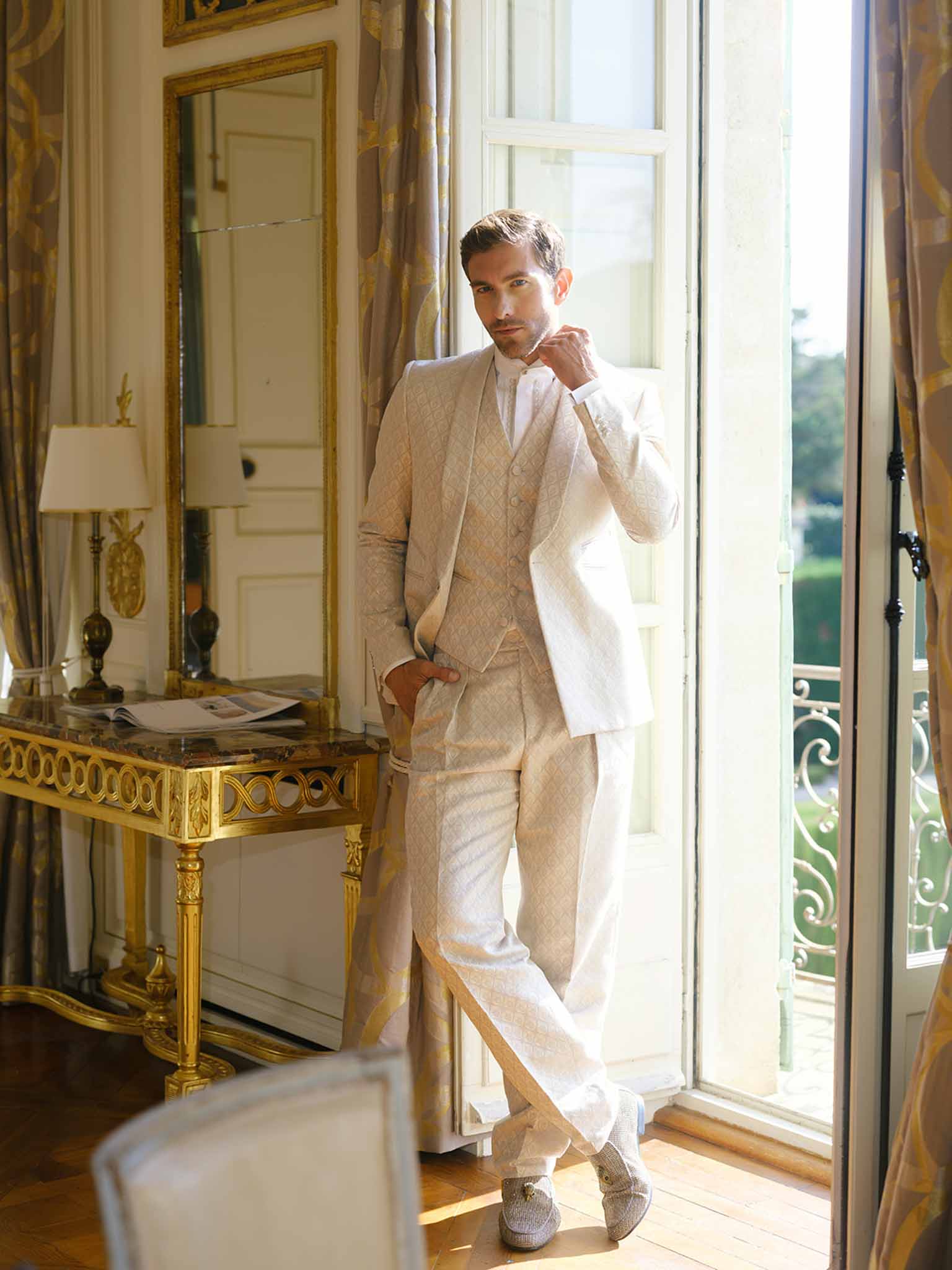 Groom adjusting tie in cream suit during getting ready session at French château interior