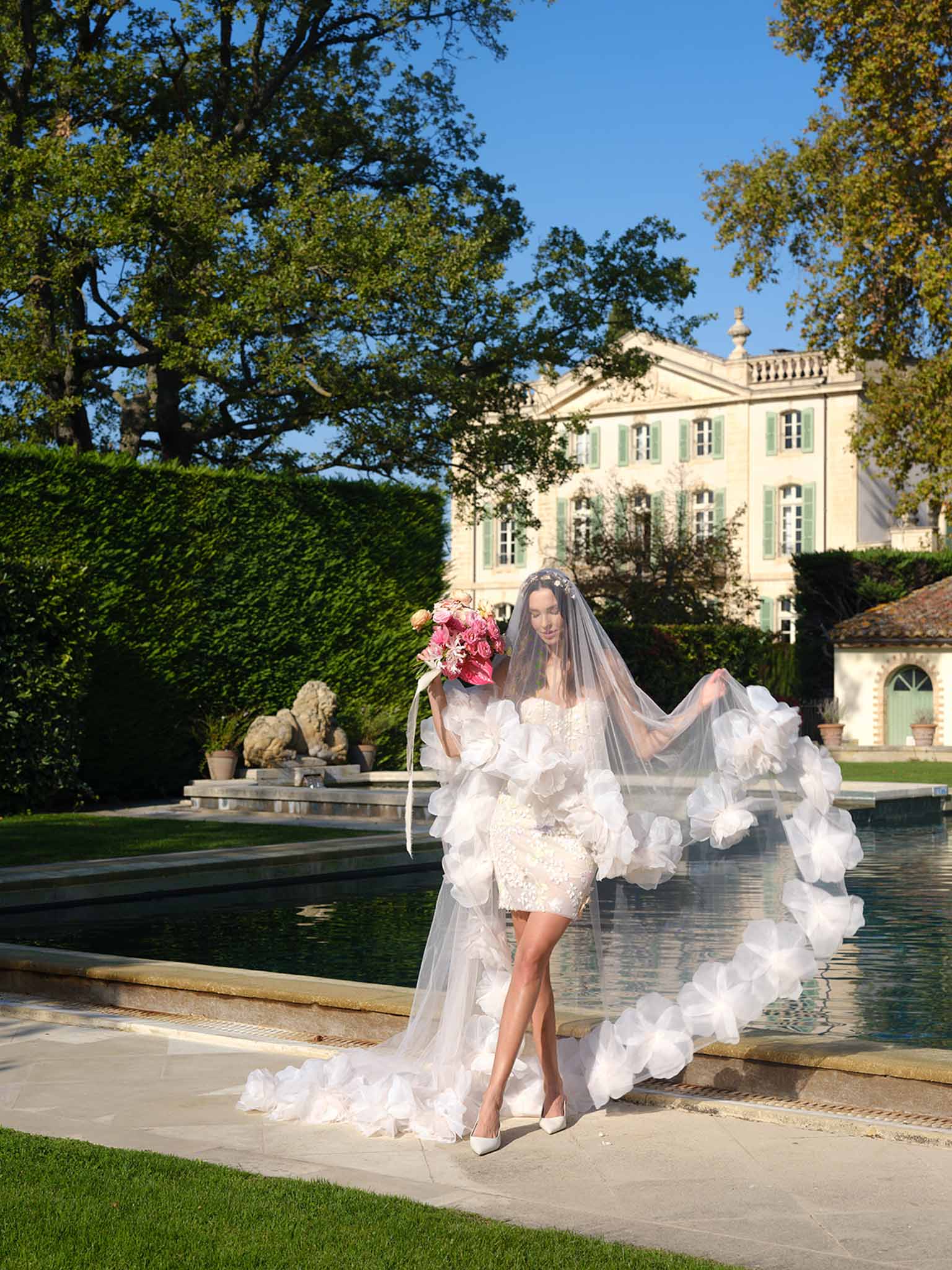 Bride in ivory lace dress with tulle train posing beside reflecting pool at elegant chateau venue