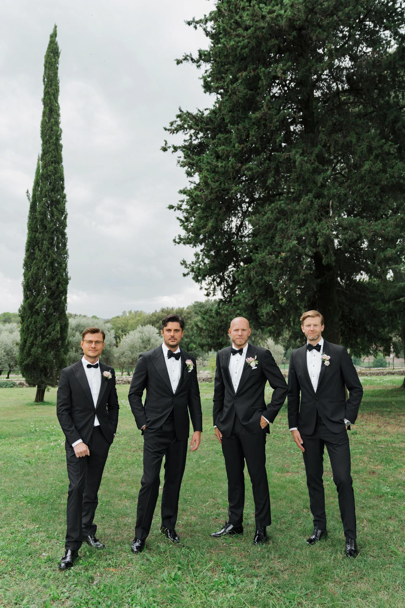 Groom and three groomsmen in matching black tuxedos with blush and white boutonnieres on estate lawn