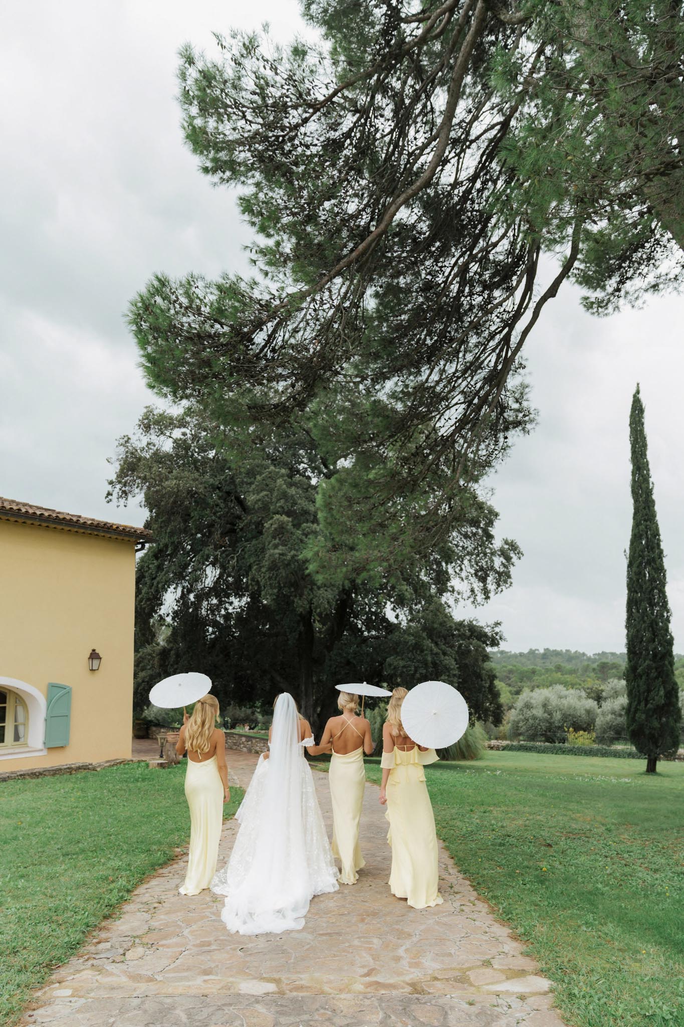 Bride and three bridesmaids in pale yellow dresses walking with white parasols at a Provencal estate
