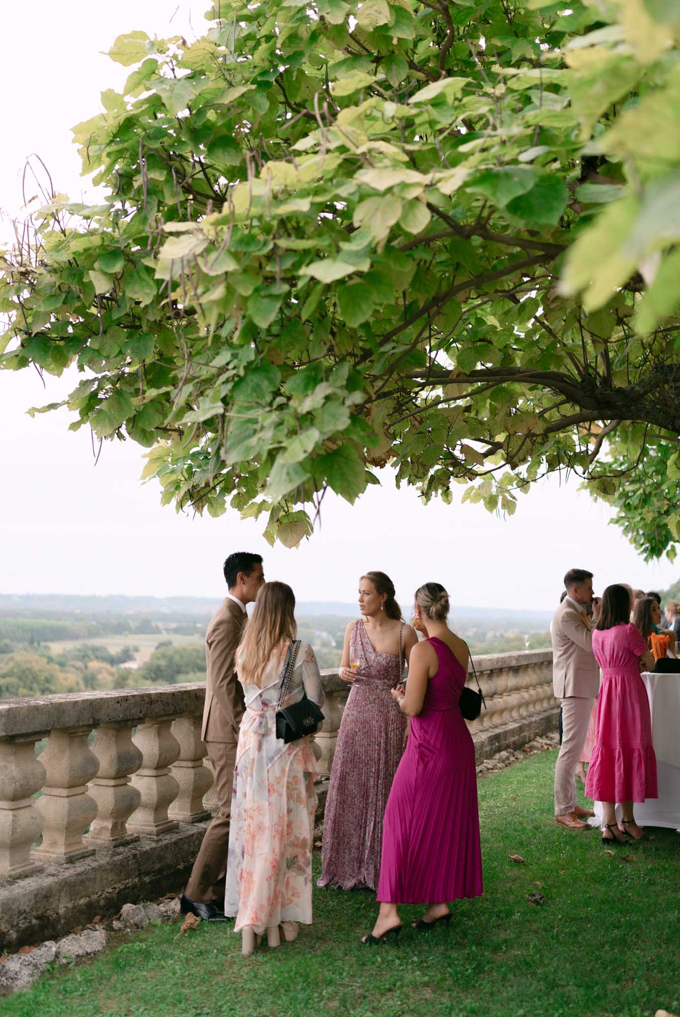 Guests in colorful dresses mingling on stone-balustraded terrace with countryside views at cocktail hour