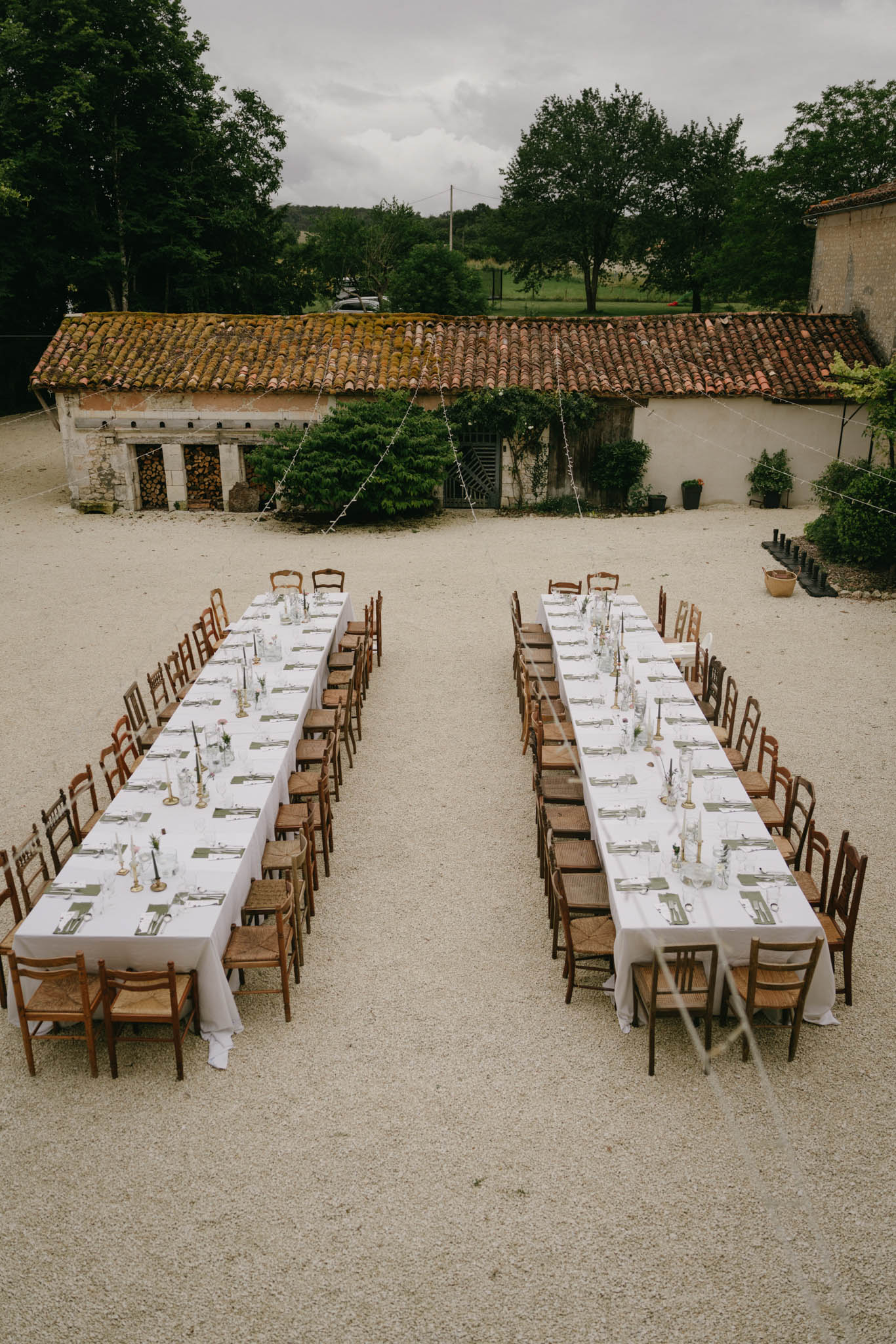 Outdoor reception with two long tables on gravel courtyard, fairy lights, vintage chairs, and stone outbuildings