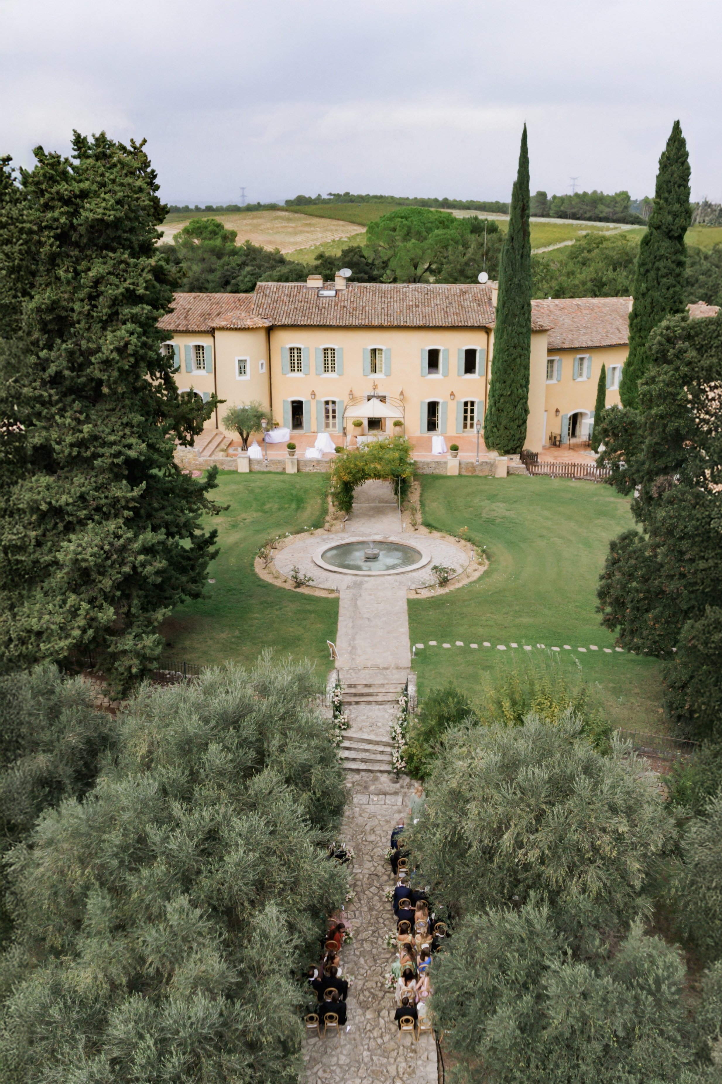 Aerial ceremony along olive tree lined path with gold chairs facing ochre Provencal mas with fountain and rose arch